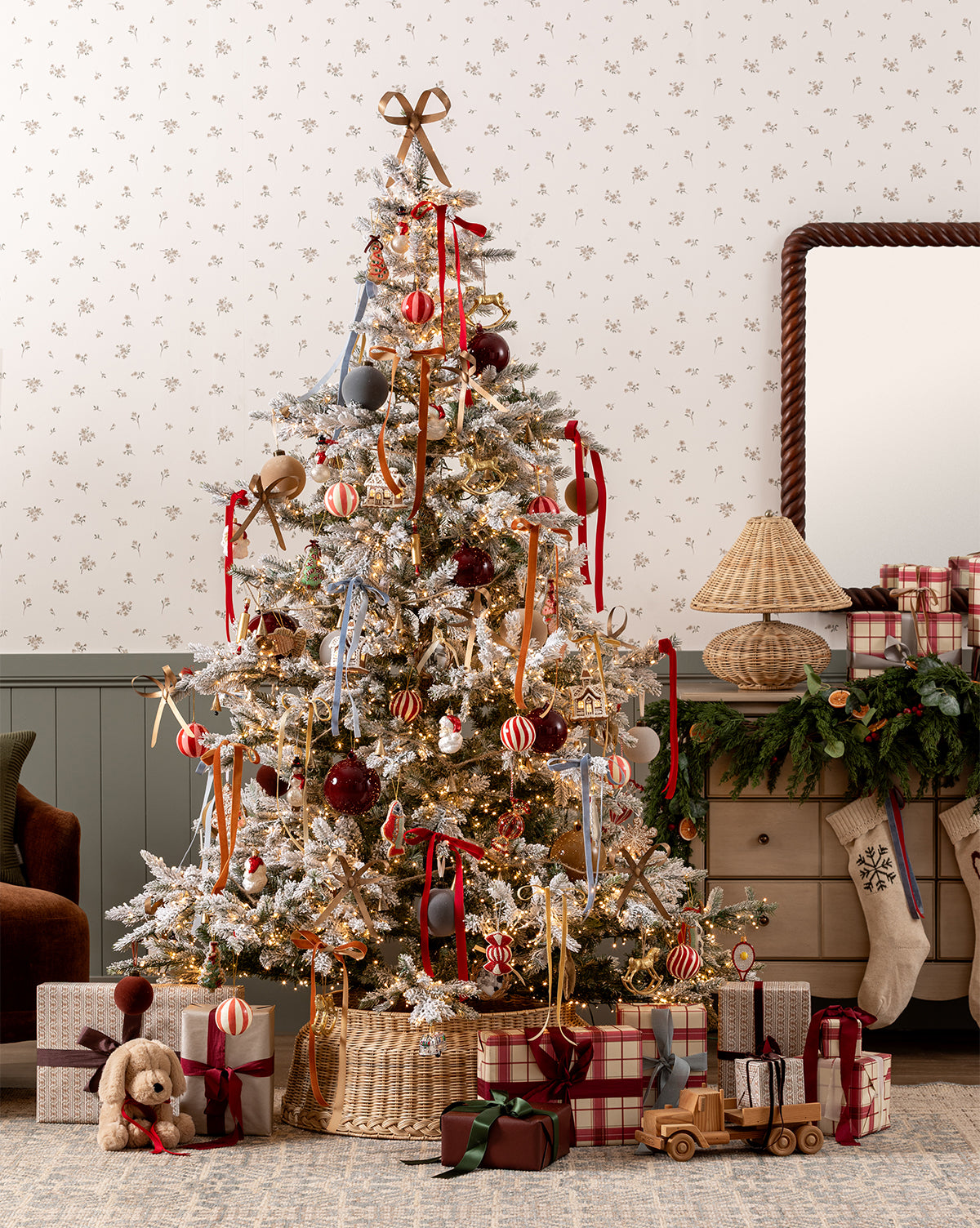 A 7' Snowy Grandis Fir Pre-Lit Faux Tree with red, gold, and striped ornaments stands in a cozy room. Wrapped gifts, a teddy bear, and a wooden toy truck are beneath it. A wreath and plaid accents decorate the background dresser, by Vendixs