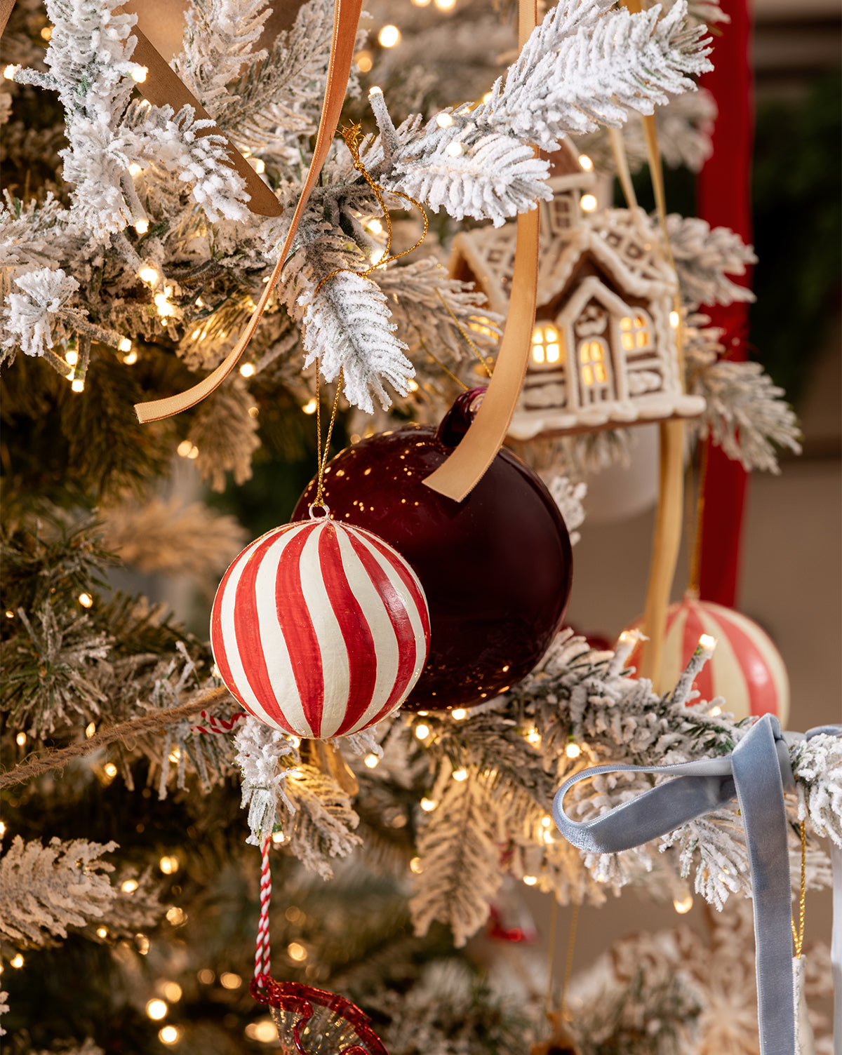A snow-frosted Christmas tree adorned with red and white striped ornaments, a maroon ball, gold ribbons, warm white string lights, a small house ornament, and 3" Hand-Painted Paper Mache Baubles with Tassel (Set of 3), by Vendixs