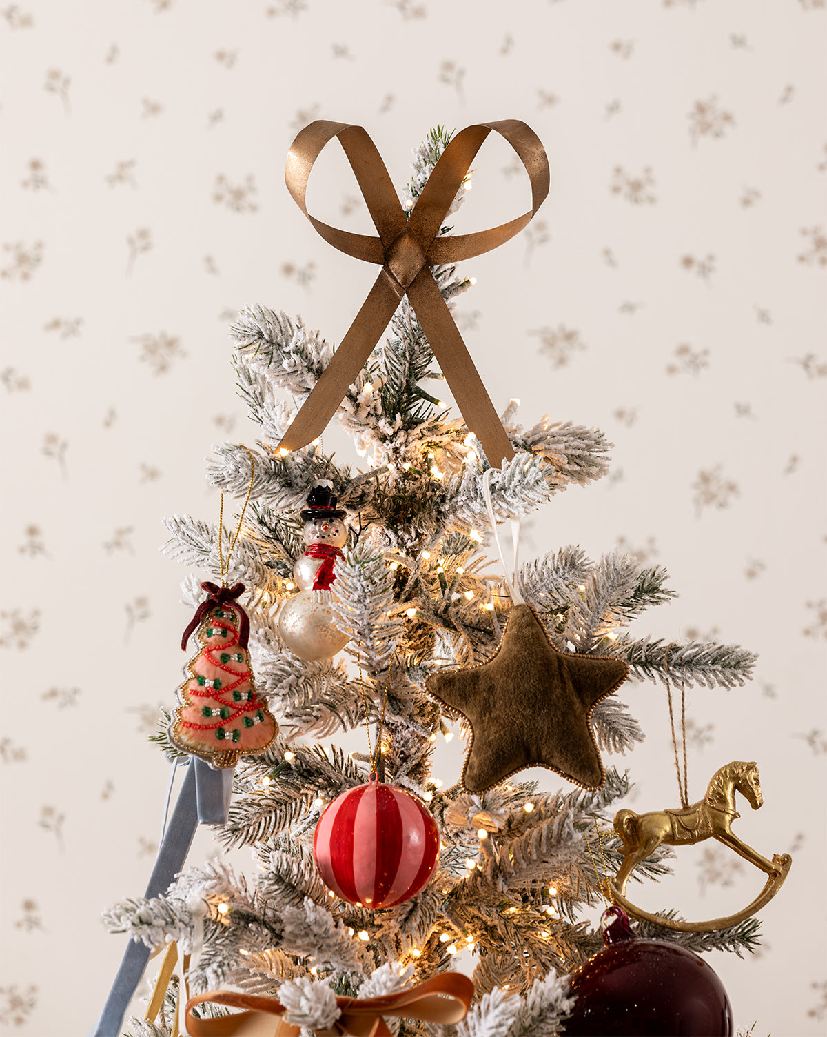A small, snow-dusted Christmas tree adorned with a Brass Bow Tree Topper, festive ornaments, and a red and white ball stands out against light floral wallpaper for a charming holiday accent, by Vendixs