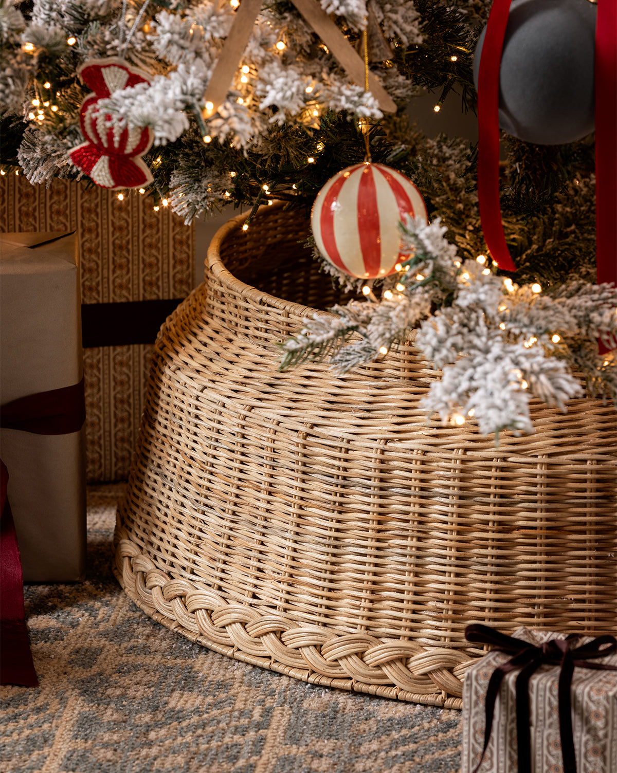 A Braided Wicker Collar surrounds the base of a Christmas tree adorned with red and white ornaments. Gifts with ribbon rest on a patterned rug, while snow-dusted branches and string lights add festive charm to the holiday decor, by Vendixs