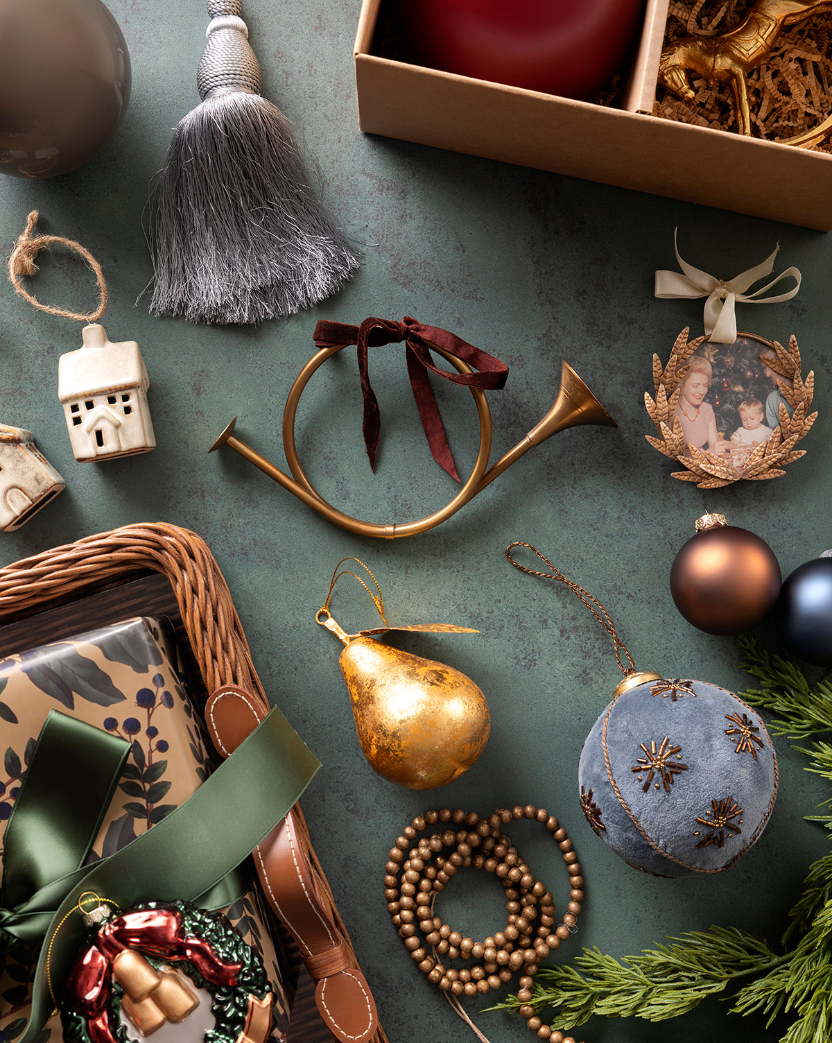 A flat lay of Christmas ornaments, including the Gilded Pear Ornament, beaded garland, brass horn, vintage ornament, baubles, framed photo, ceramic house, tassel, wrapped gifts, and greenery on a green tabletop, by Vendixs
