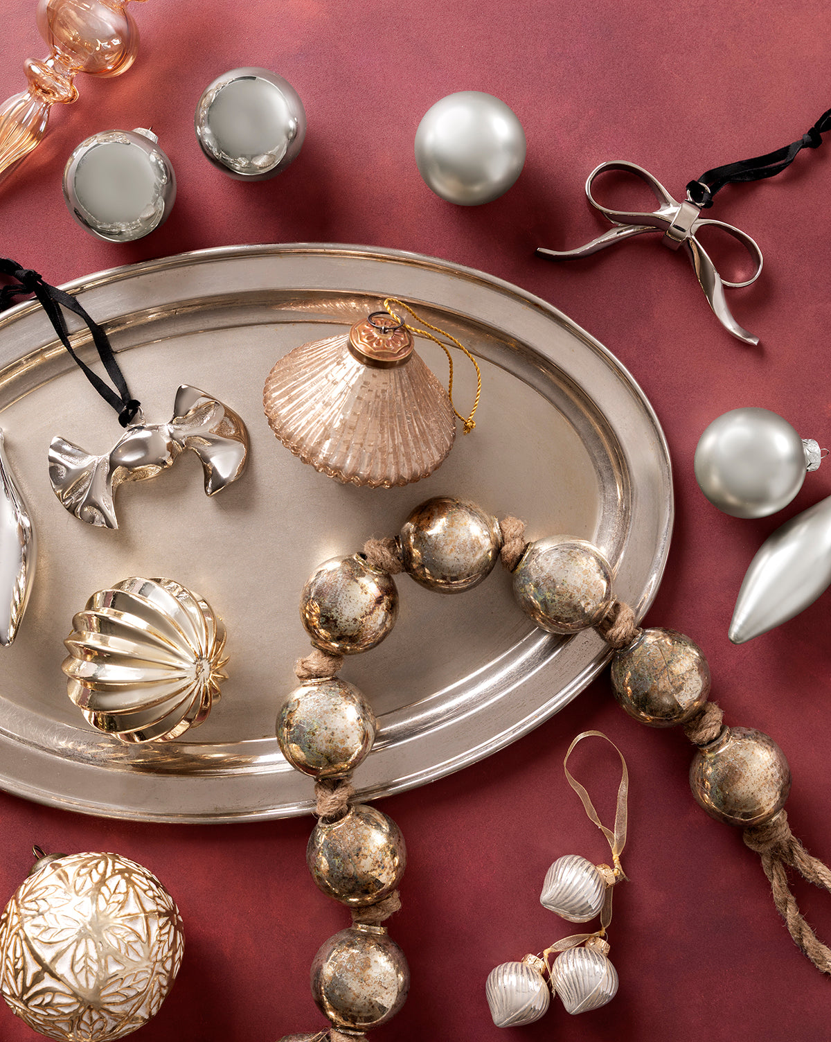 A silver tray with gold and metallic ornaments, including bows, shells, balls, and beaded garland—plus Piper Ornaments—displayed on a red background with gunmetal Christmas tree decor scattered around, by Vendixs
