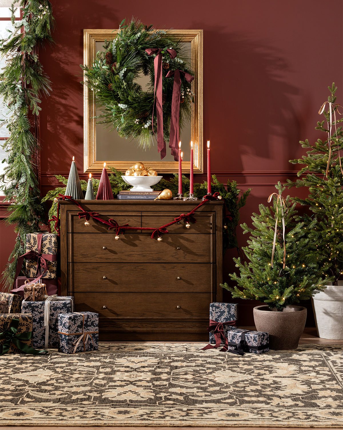 A festive room features a wooden dresser with candles, gold ornaments, and decorative trees. Above, a mirror displays the 24" Pinecone & Berries Faux Wreath. Wrapped presents and seasonal decor sit by the potted Christmas tree, by Vendixs
