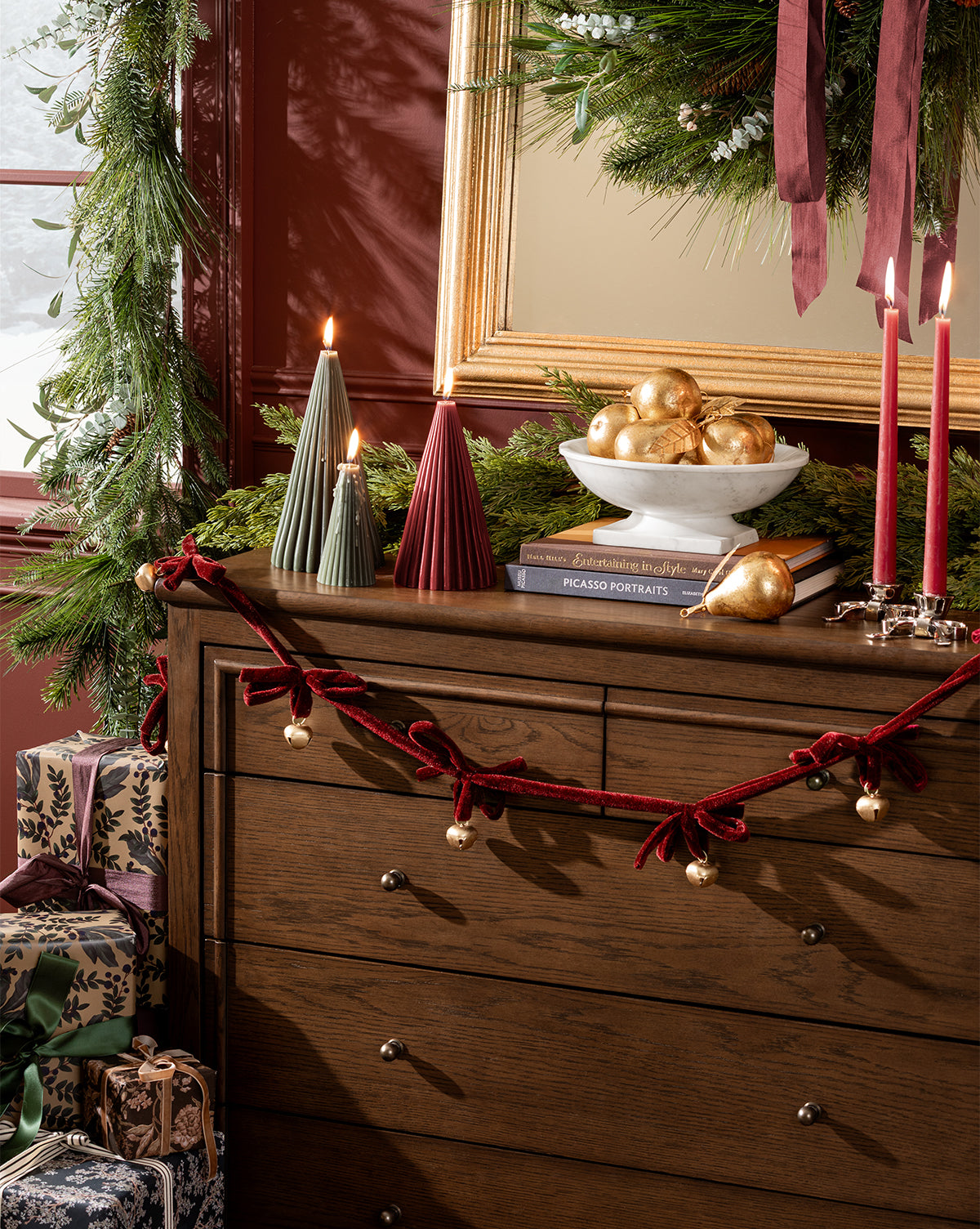 A wooden dresser is decorated for the holidays with green garland, candles, wrapped gifts, a bowl of vintage ornaments, gold patina accents, a red velvet bell garland, and features the Gilded Pear Ornament. A large wreath hangs above by Vendixs