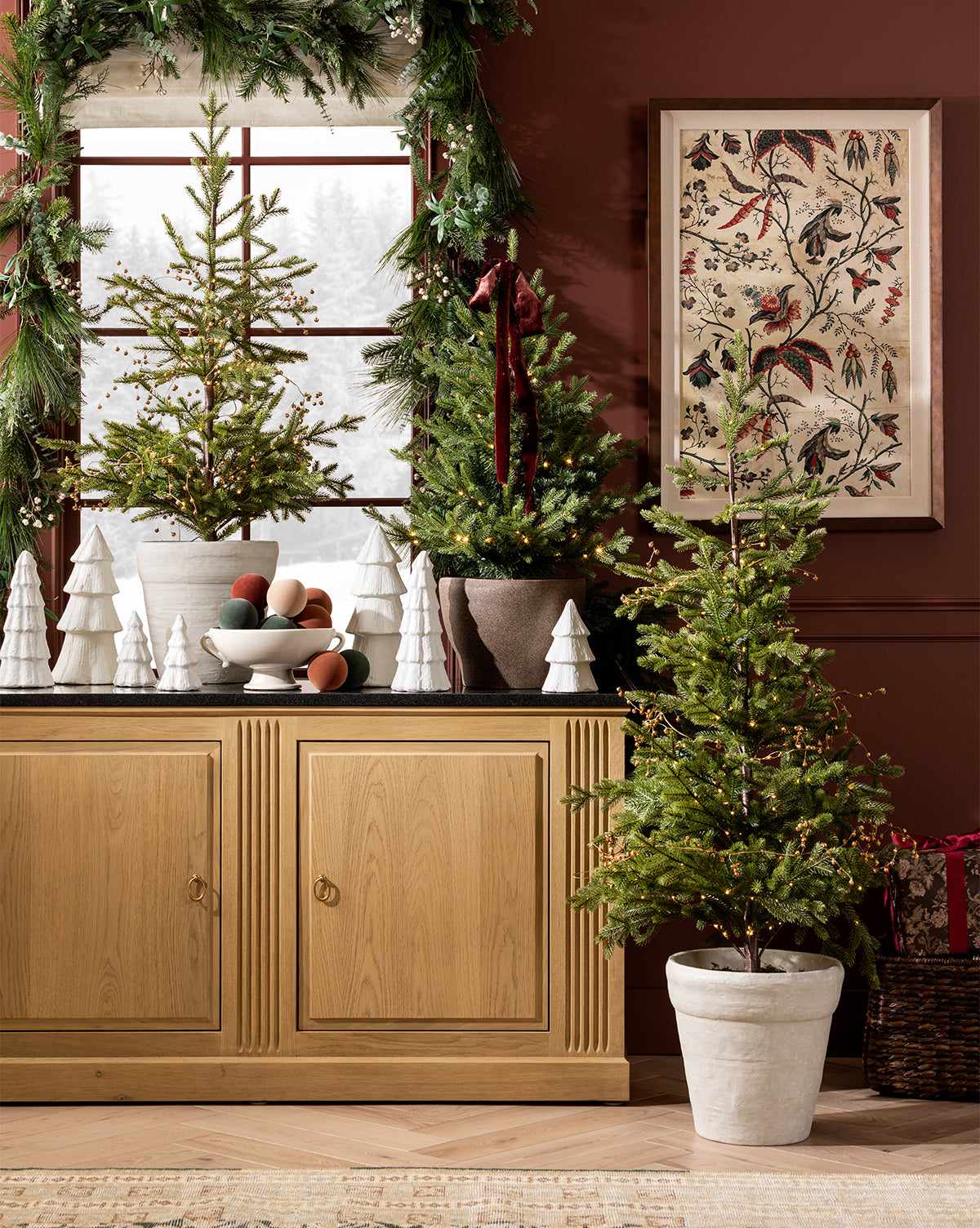 A holiday scene features Confectioners Paper Mache Tree figurines in white pots on a wooden sideboard, with a bowl of ornaments, garland around the window, and bird art decorating a maroon wall, by Vendixs