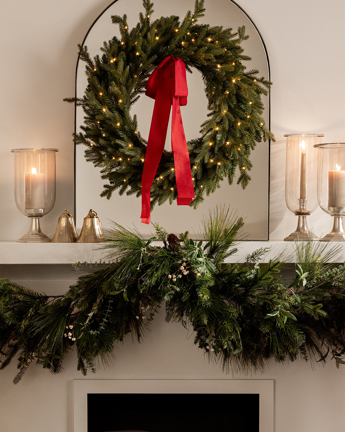 A festive mantel is adorned with a vintage-inspired green wreath with a red ribbon, two lit candles in glass holders, Polished Brass Bell ornaments, and a pine garland for charming holiday décor, by Vendixs