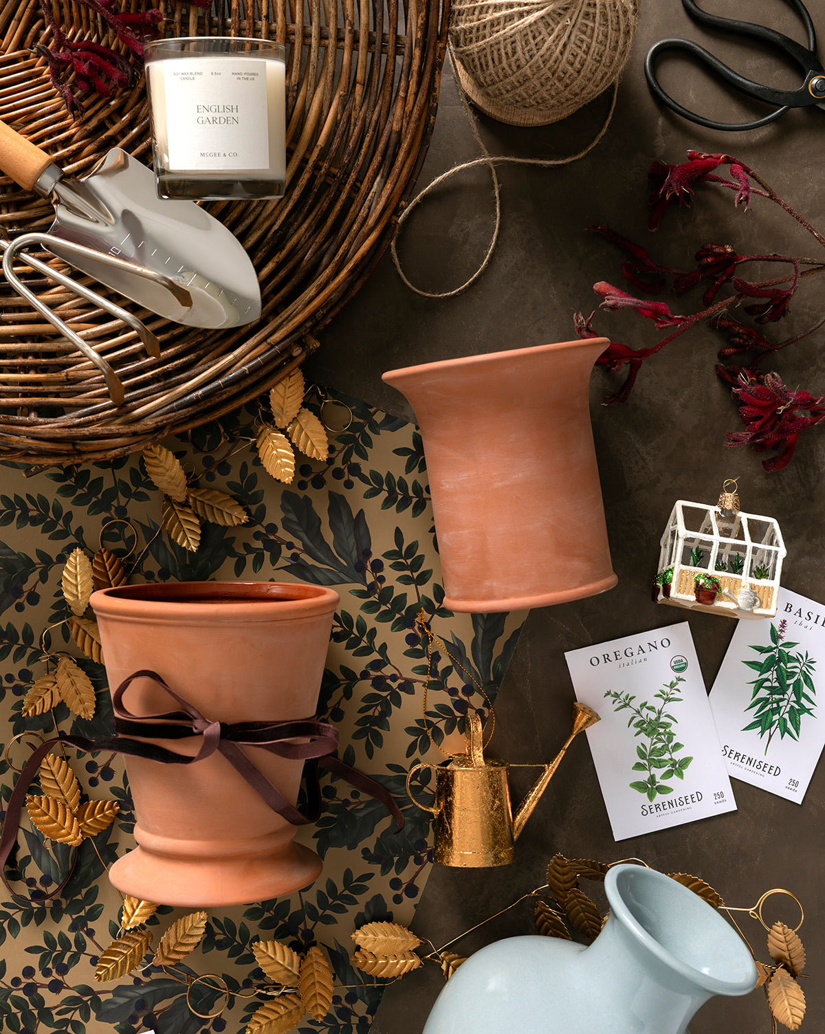A flat lay of gardening items, including terracotta pots, a gold Watering Can Ornament with a copper finish, a white trowel, scissors, seed packets, twine, a candle, and leafy decorative paper on a brown surface, by Vendixs