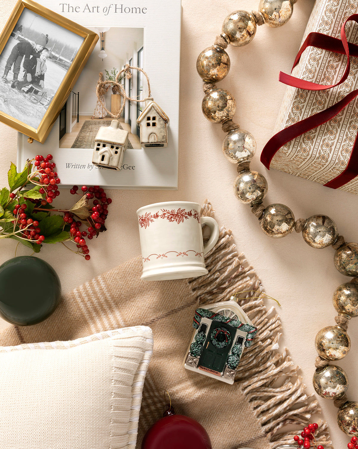 A cozy flat lay with a cream blanket, pillow, festive mug, framed photo, berries, candles, metallic garland, wrapped gift with ribbon, the Glazed Porcelain Houses (Set of 2) as festive accents, and the book “The Art of Home”, by Vendixs