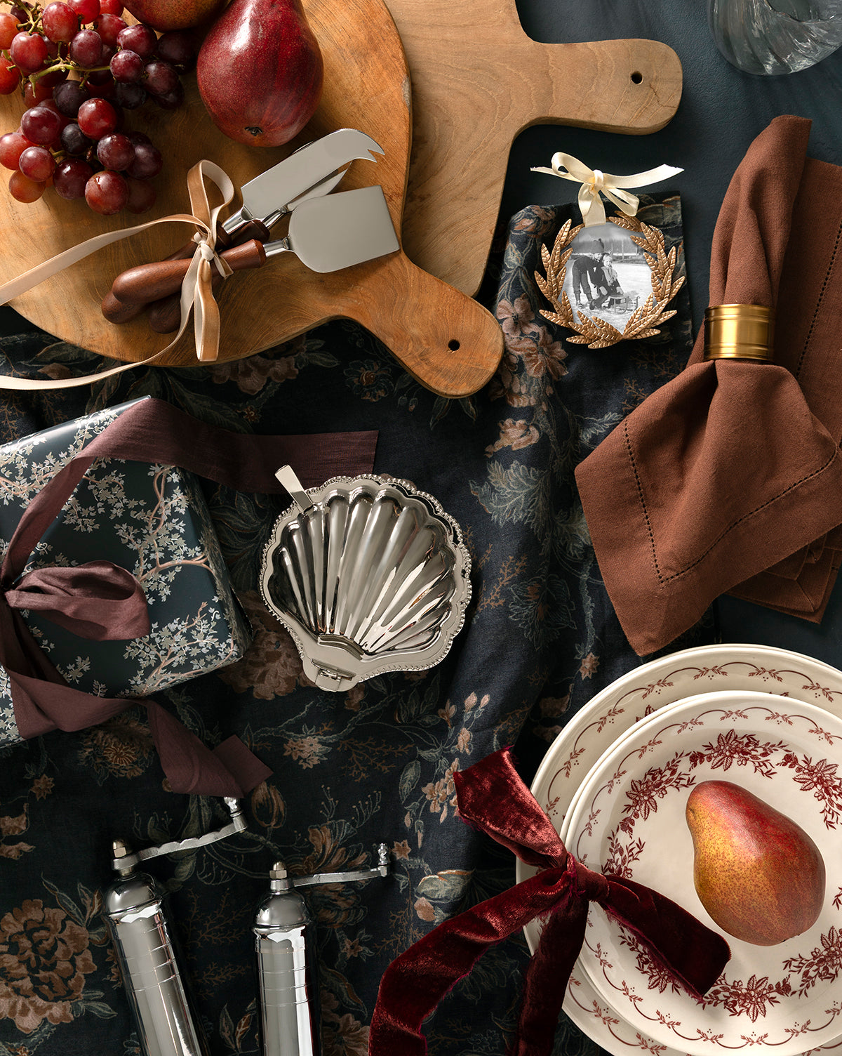 A flat lay of elegant holiday tableware featuring plates with red patterns, a pear, ribbon-tied gifts, brown napkins with gold rings, grapes, the Fromagerie Cheese Knives (Set of 3), and a festive ornament on a dark floral tablecloth, by Vendixs