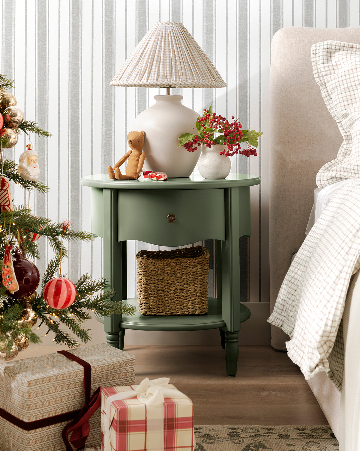 A green round nightstand with a lamp, teddy bear, and festive decor—including the Lyra Vase—sits beside a beige bed. A basket is on the lower shelf. A Christmas tree and wrapped gifts stand in front of striped wallpaper, by Vendixs