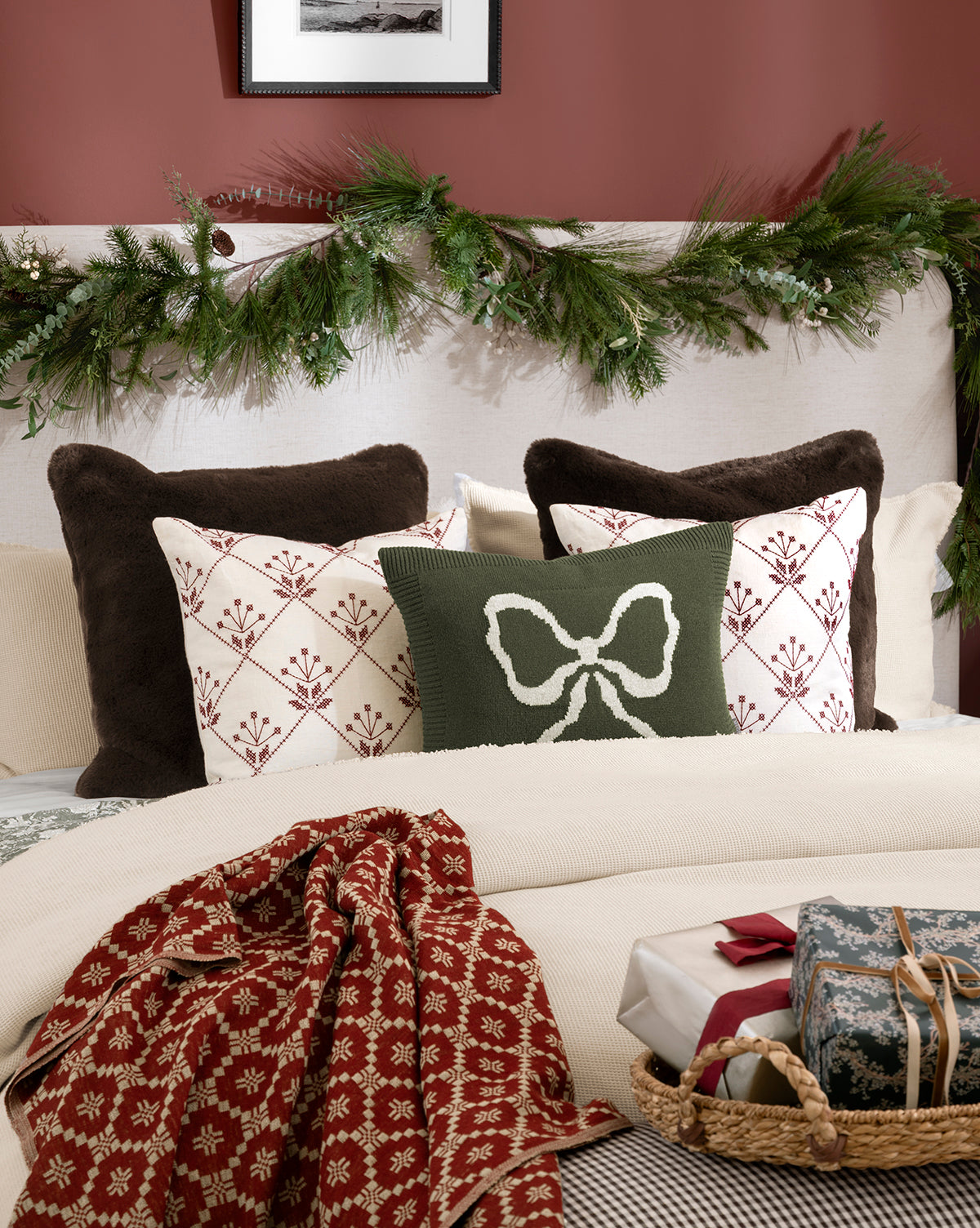 A bed with patterned pillows, featuring the Bella Pillow Cover and a green pillow with a white bow, is styled with a red and white throw. Holiday touches include an evergreen garland on the headboard and wrapped gifts at the foot of the bed, by Vendixs