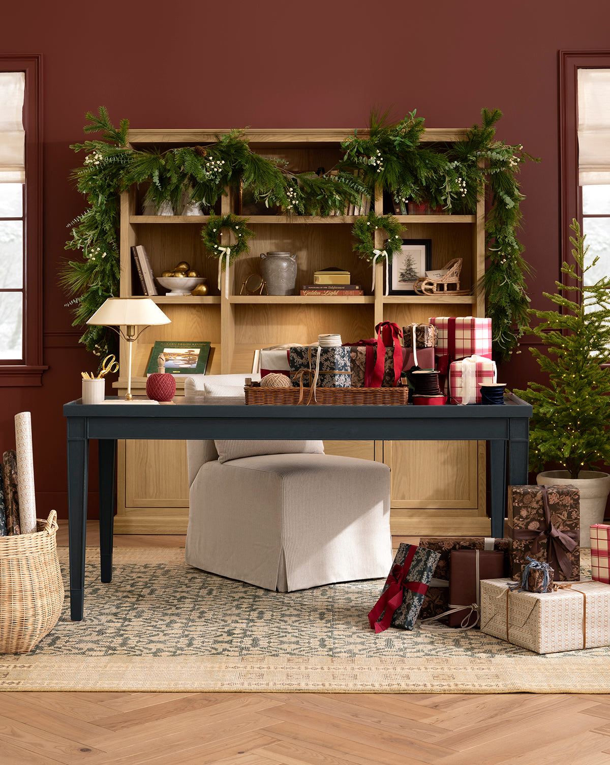 A festive home office featuring the 6' Pinecone & Berries Faux Garland on the bookshelf, a navy blue desk, beige chair, wrapped gifts, basket, small decorated Christmas tree, and holiday décor in warm lighting, by Vendixs