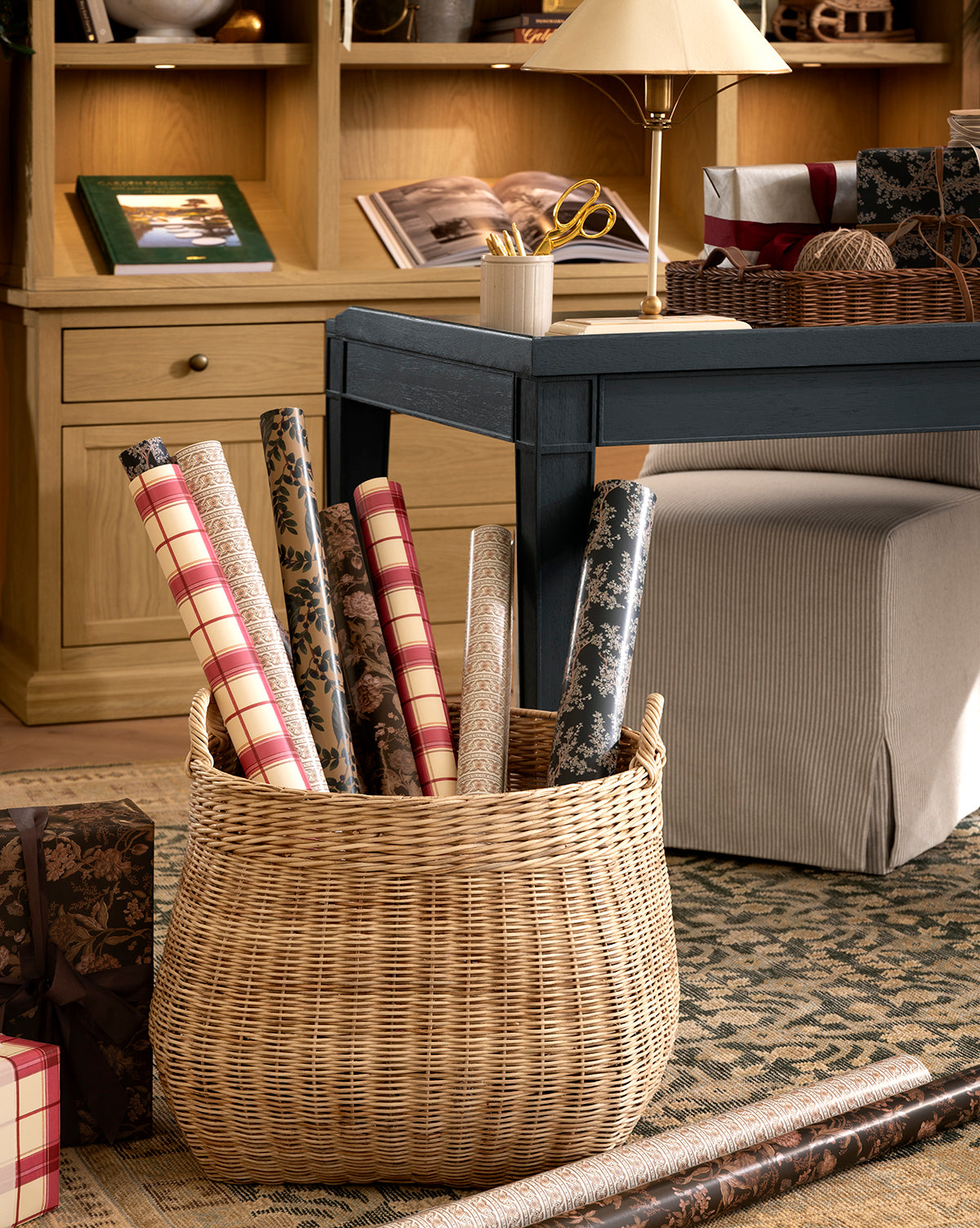 A wicker basket filled with Antigone Branch Gift Wrap rolls rests on a patterned rug in a cozy room with a desk, books, lamp, and wrapped gifts in the background, by Vendixs