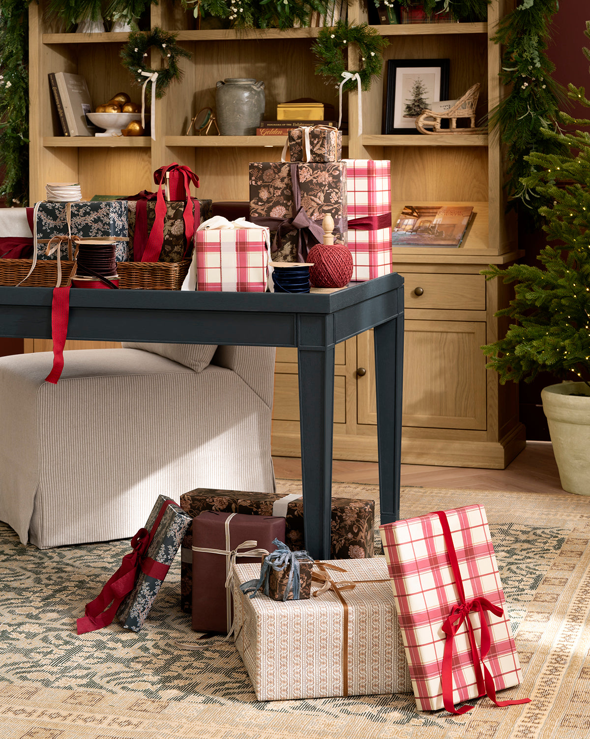 A festive room with a dark-covered table surrounded by gifts wrapped in Highland Plaid Gift Wrap and others, red and pink ribbons, bookshelves adorned with garland, and a Christmas tree in the corner, by Vendixs