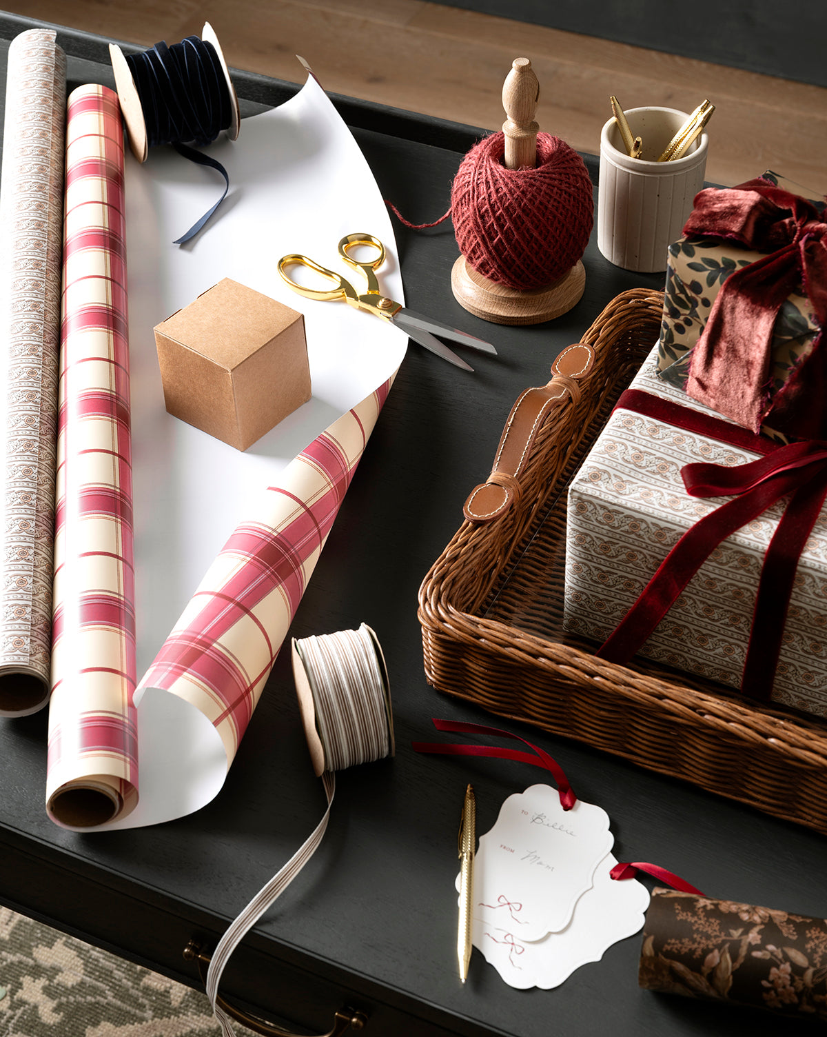 A table with gift wrapping supplies: patterned paper, scissors, 3/8" Velvet Ribbon with Woven Edge, twine, a pen, a gift tag, a plain box, and a wicker basket holding wrapped gifts with bows for festive holiday décor, by Vendixs