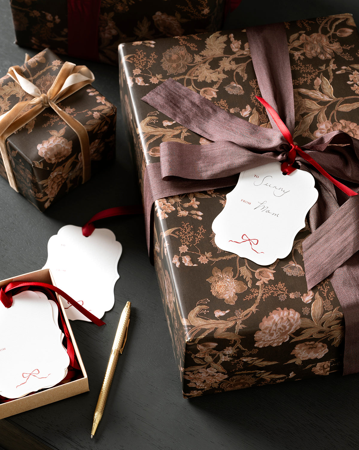 Several gifts wrapped in elegant brown floral paper with wide velvet ribbons and white Paperie Gift Tags (Set of 24) rest on a dark surface. A gold pen and extra blank tags are nearby, ready for holiday gifting, by Vendixs