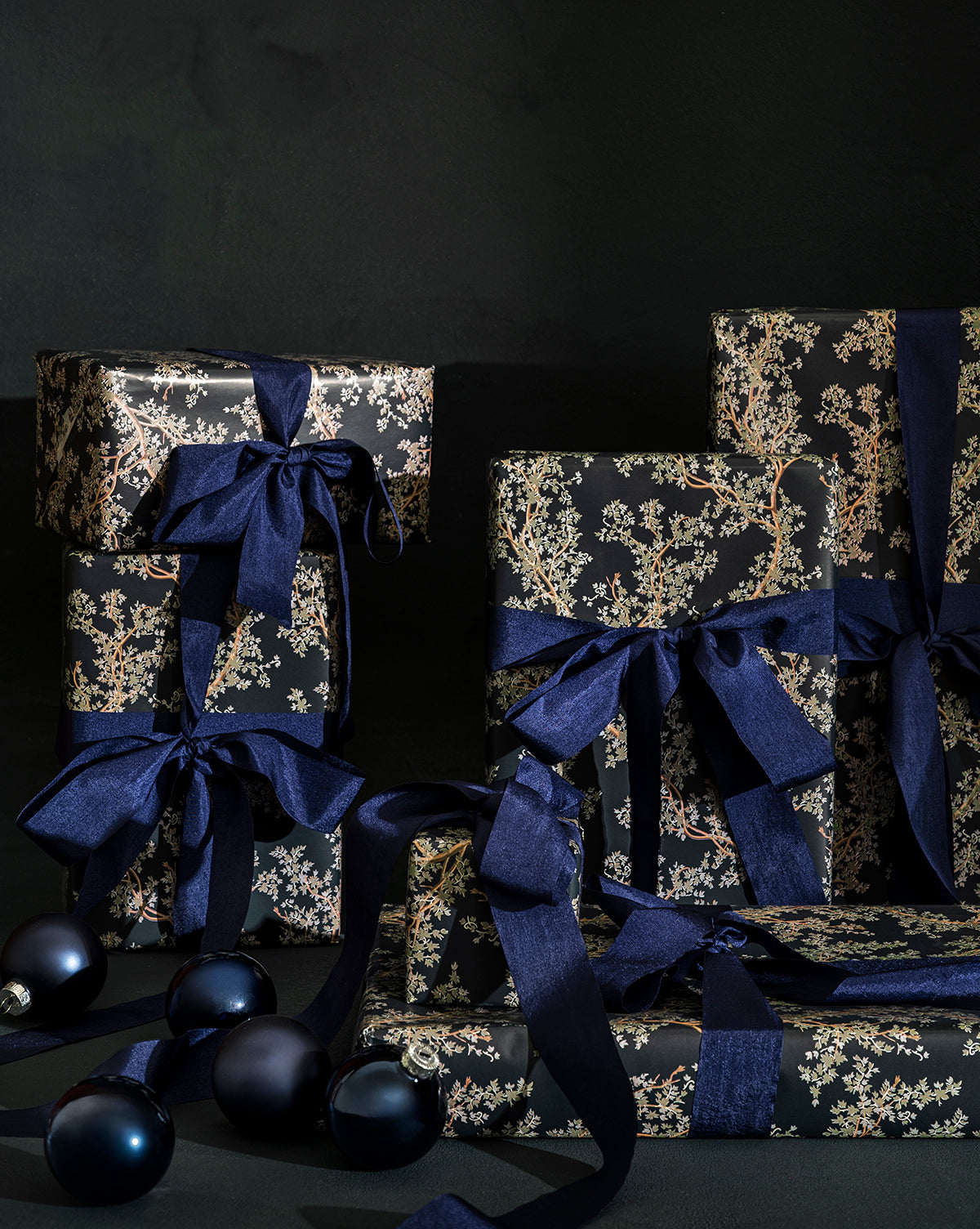 Several gifts wrapped in Antigone Branch Gift Wrap with navy blue ribbons are stacked together, with dark blue ornaments scattered in front, all set against a dark background for an elegant holiday gifting display, by Vendixs