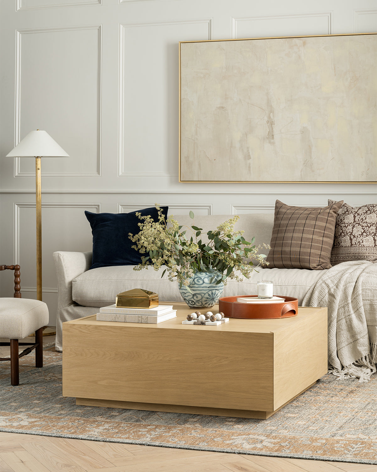 A modern living room features a light sofa, dark plaid pillows, beige blanket, Bentley Coffee Table with books, decor, and a vase of greenery, a floor lamp, accent chair, and abstract wall art by Vendixs