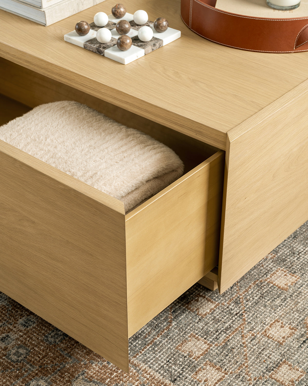 A light wood drawer of the Bentley Coffee Table is partially open, showing a folded beige towel inside. The table sits on a patterned rug with decorative objects and books neatly arranged on top, by Vendixs