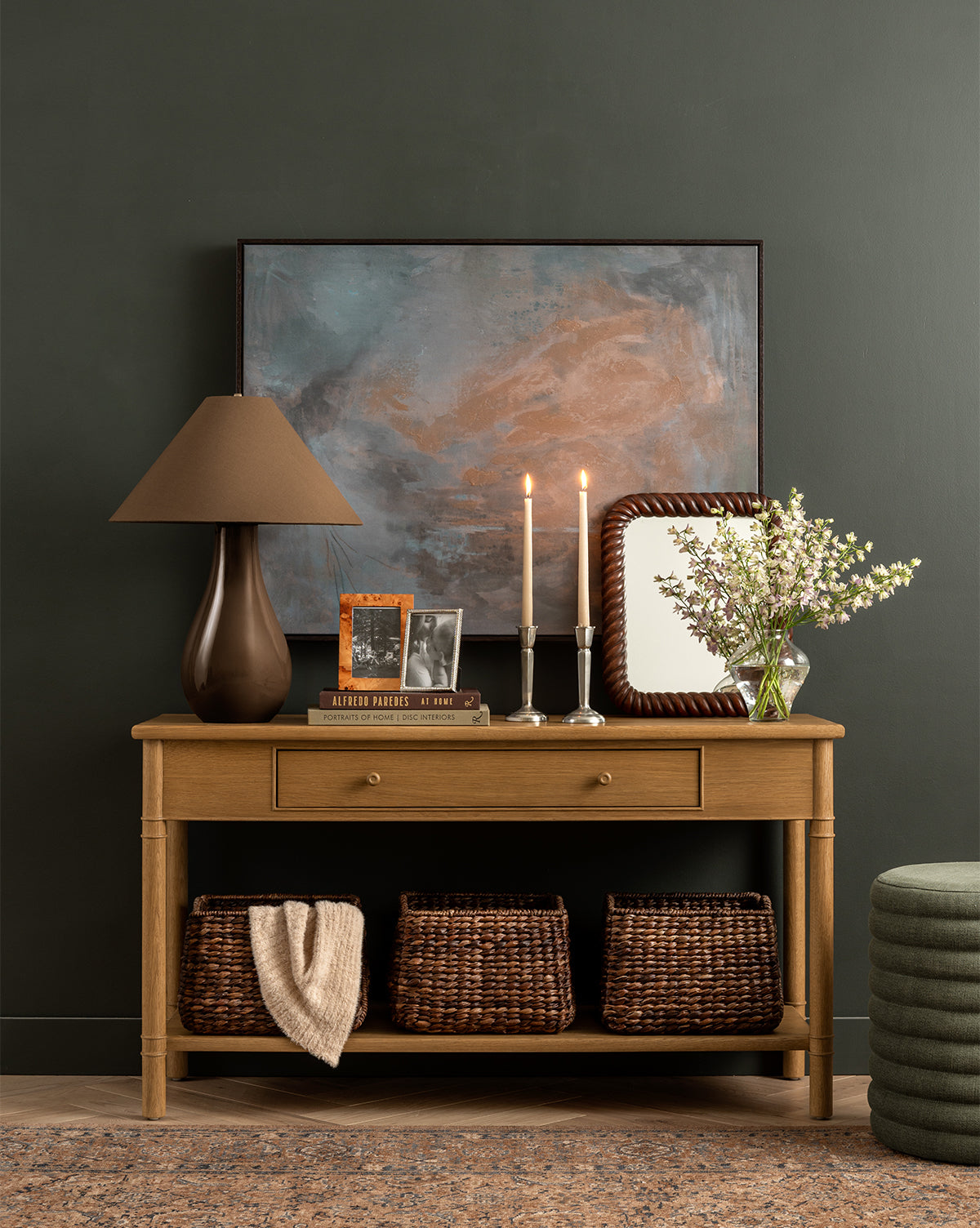 The Hindley Console stands against a dark wall displaying a brown lamp, framed photos, candles, mirror, vase with white flowers, and books. Its lower shelf holds three woven baskets and a folded cloth, making it perfect for any entryway, by McGee & Co.