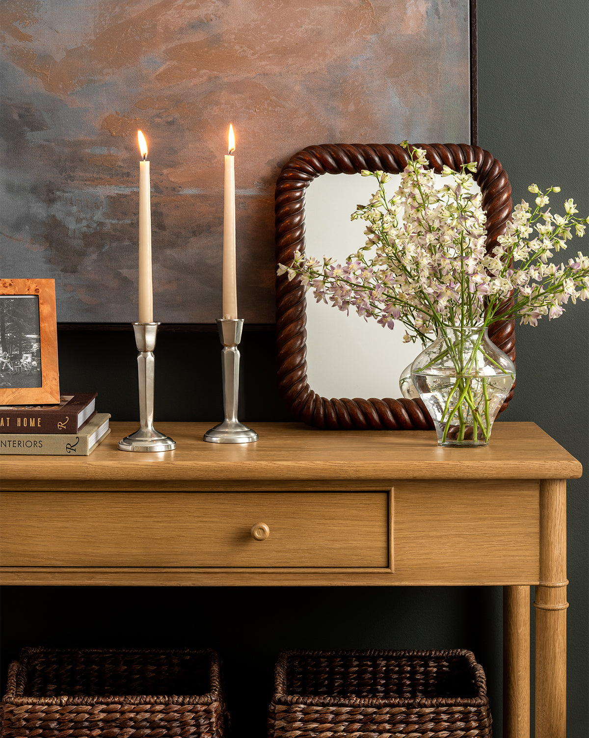 The Hindley Console holds two lit candles in silver holders, a small stack of books, a framed photo, a vase of white flowers, and a braided-frame mirror against a muted wall. Two baskets are placed underneath this entryway console, by Vendixs