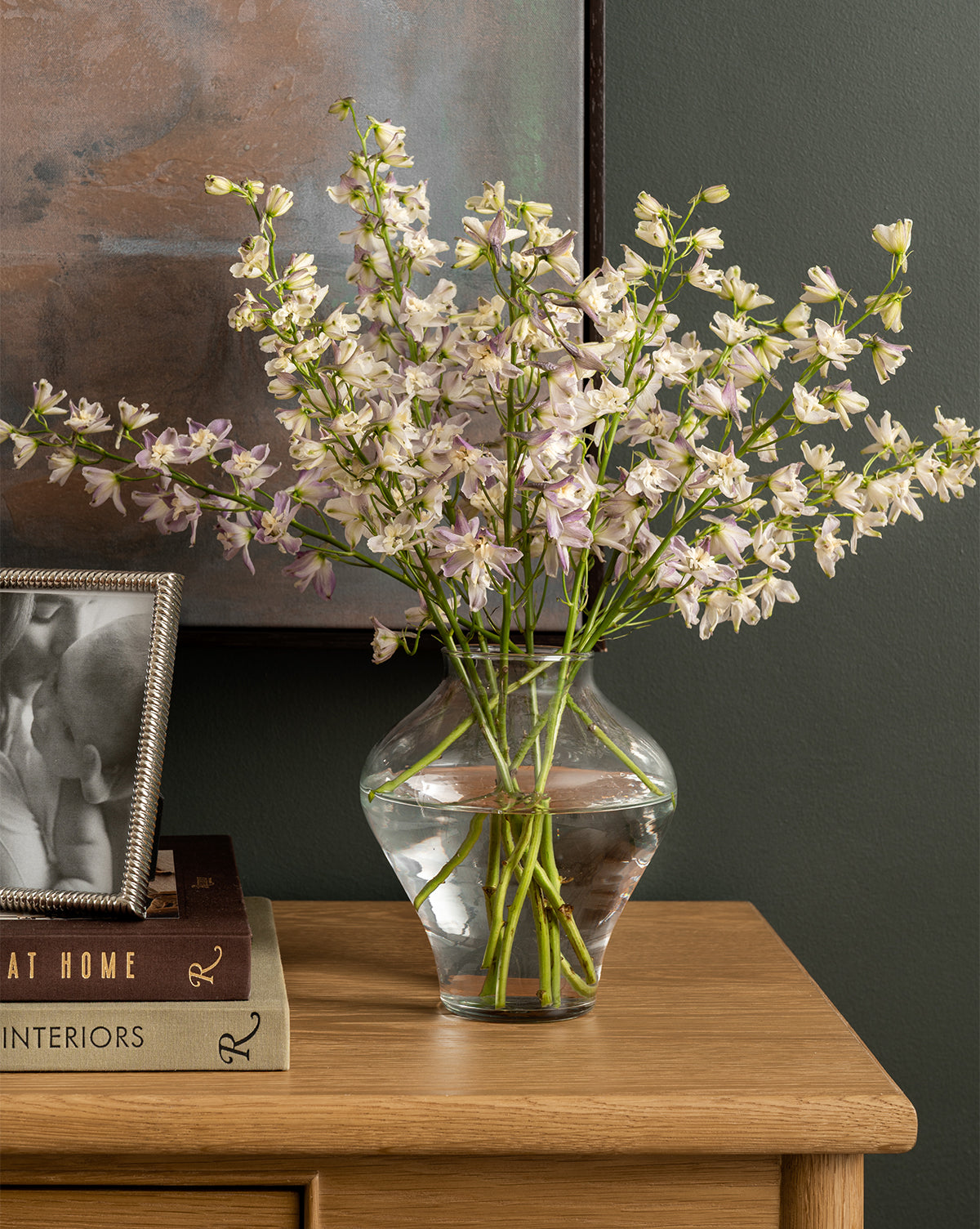 The Allaire Glass Vase, filled with water and delicate white and pale purple flowers, sits on a wooden table beside a framed photo and stacked books beneath a wall painting, by Vendixs