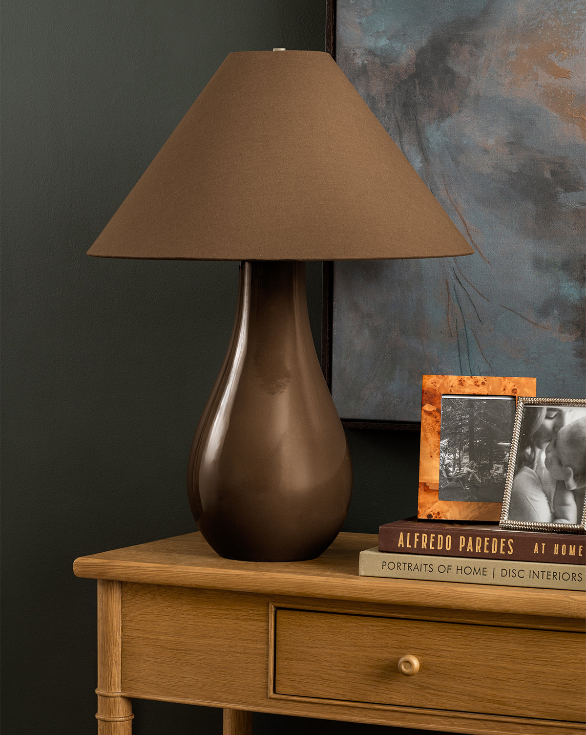 The Irving Table Lamp, featuring a sculptural brown base and wide fabric shade, sits on a wooden desk next to books and framed photos, set against a dark wall with part of a painting in view, by Vendixs