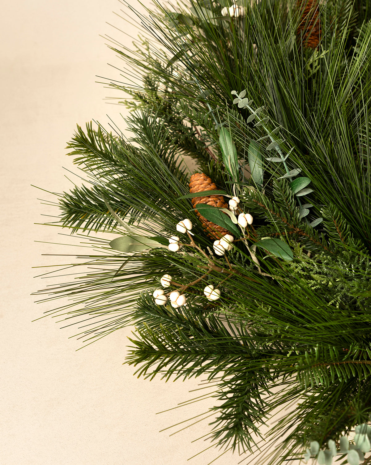 The 24" Pinecone & Berries Faux Wreath features pine branches, green foliage, white berries, and a pinecone—a charming seasonal accent for your home, displayed here against a light beige background, by Vendixs