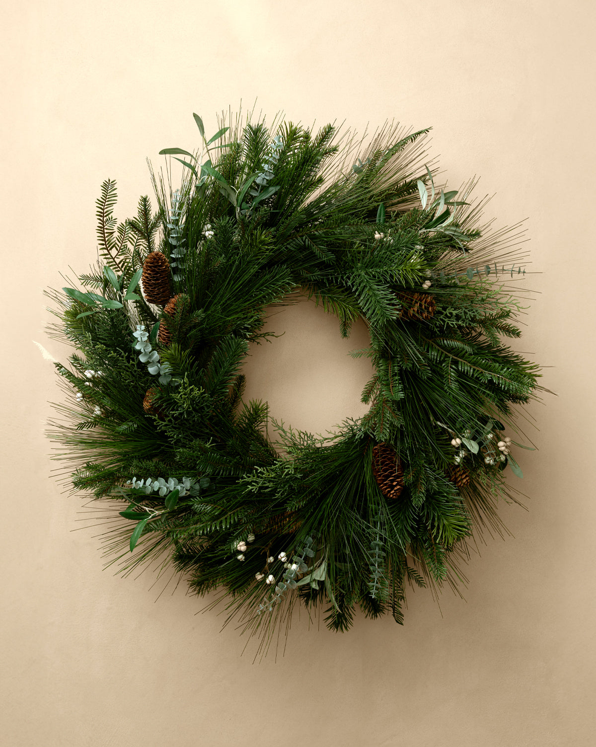 The 24" Pinecone & Berries Faux Wreath, featuring pine and evergreen branches with pinecones and white berries, adds festive charm to a plain beige wall, by Vendixs