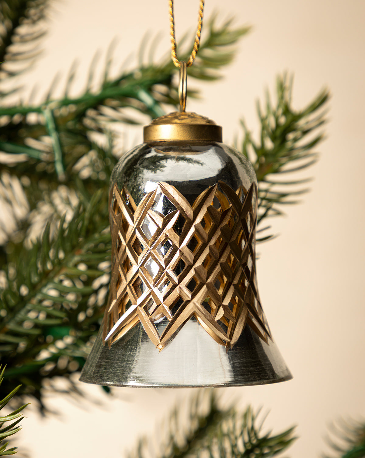 The Hand-Painted Mercury Glass Bell Ornament, in silver and gold with a detailed geometric design, hangs from green pine needles—an ideal touch for festive holiday décor, by Vendixs