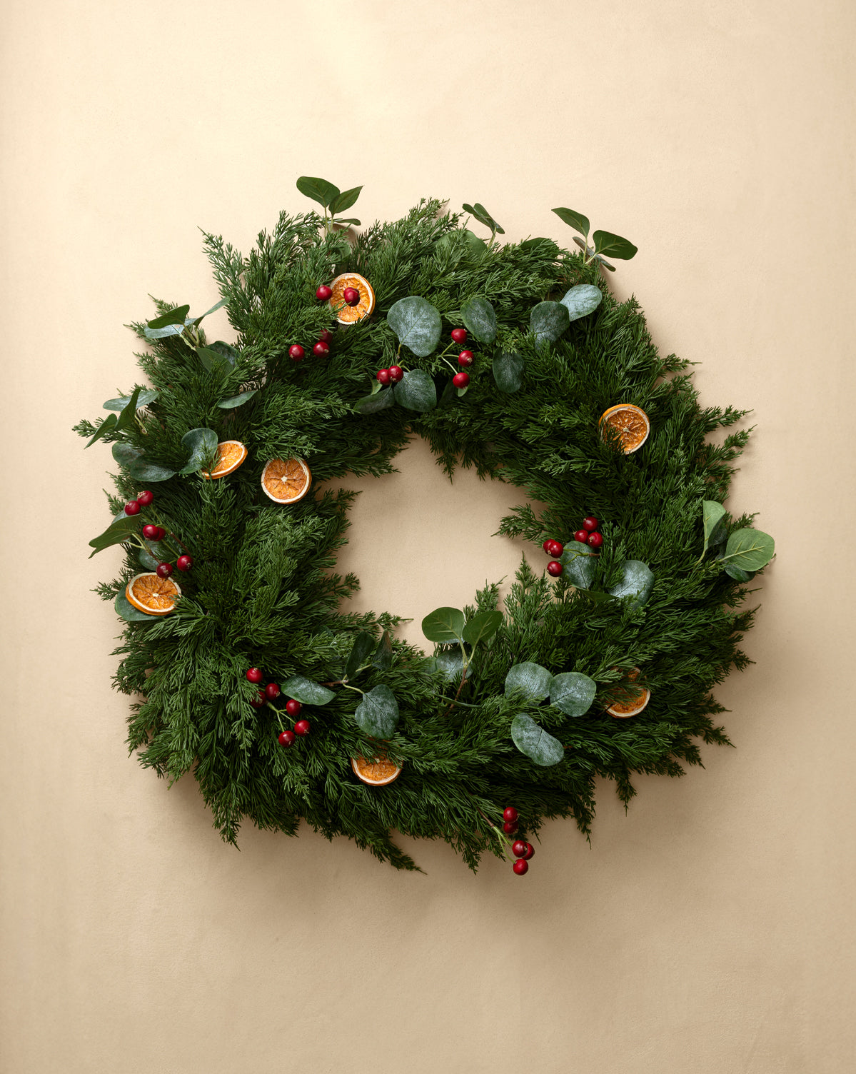 A 30" Orange & Eucalyptus Faux Wreath, adorned with red berries and dried orange slices, hangs on a plain beige wall, by Vendixs
