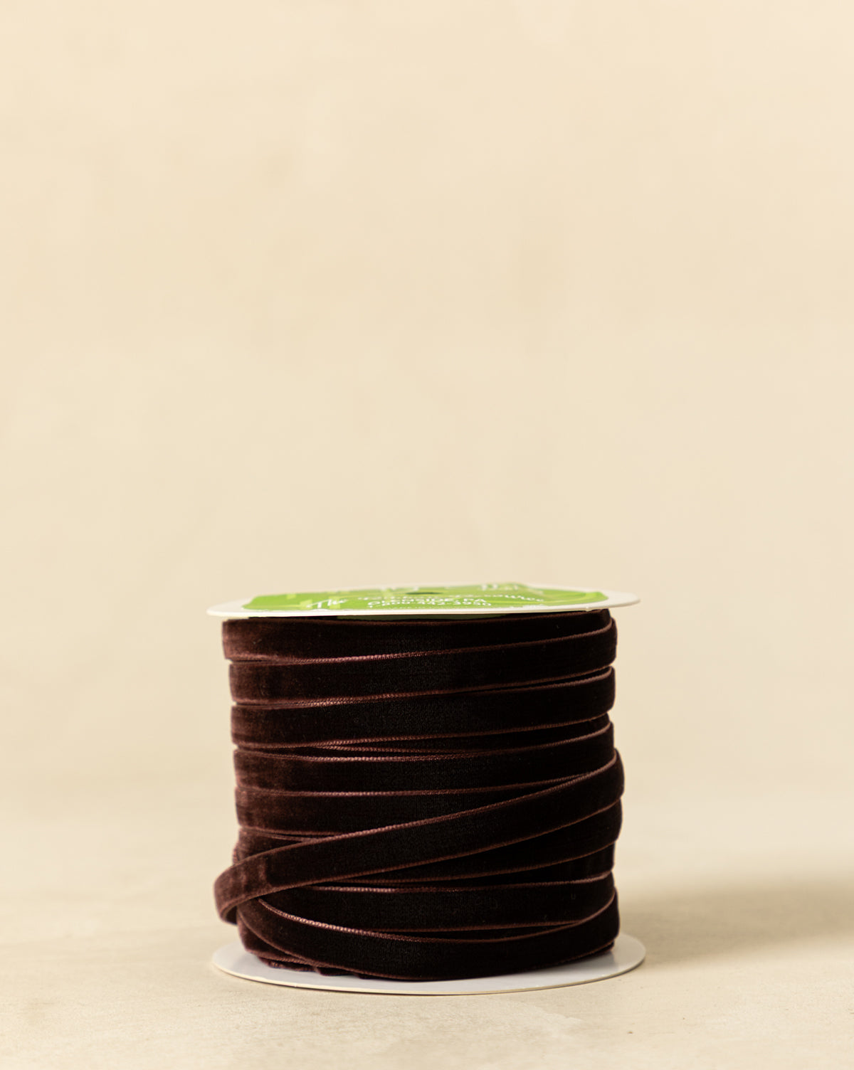 A spool of 3/8" Velvet Ribbon with Woven Edge in dark brown, featuring a white and green label, rests on a light beige surface against a plain beige background, by Vendixs