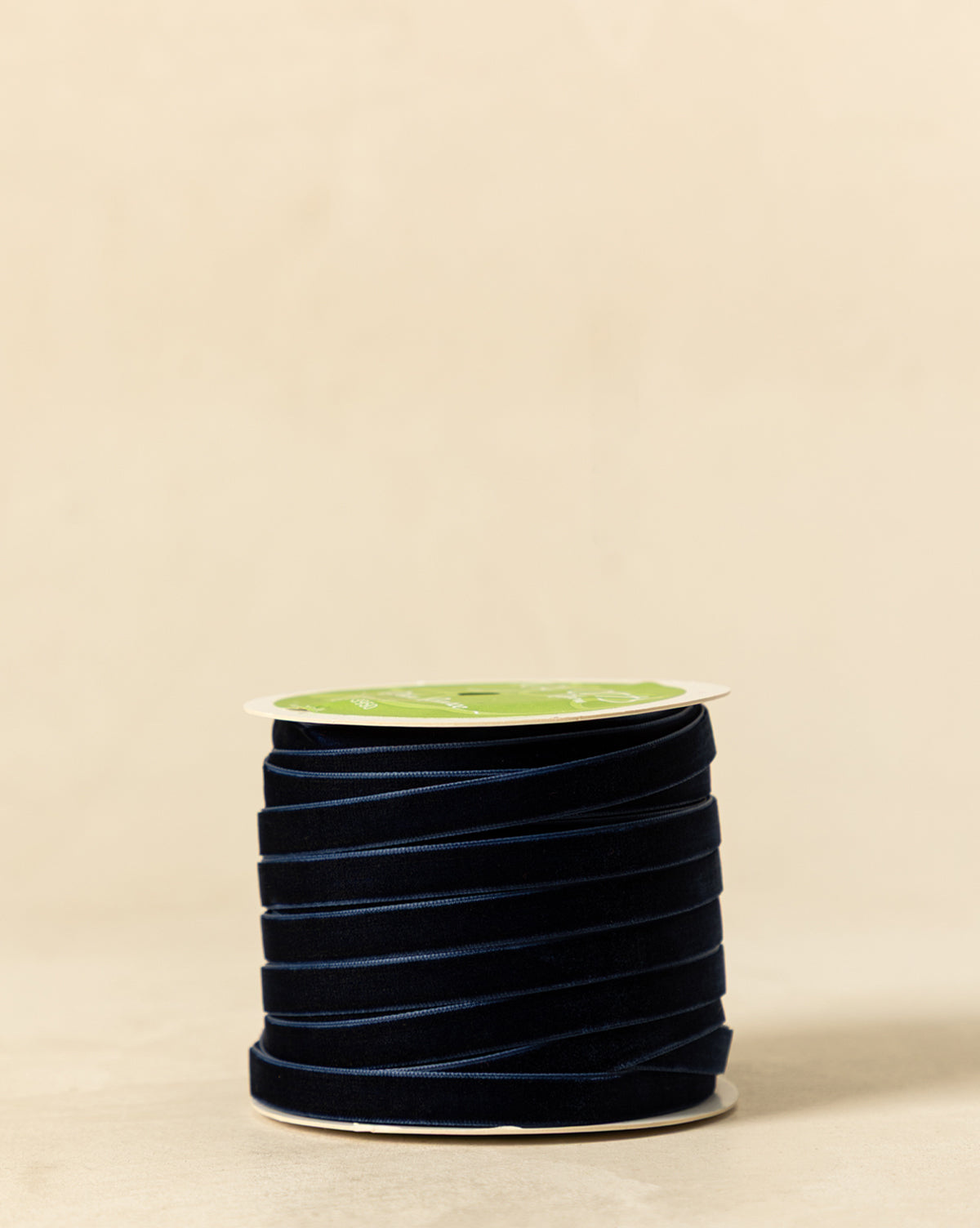 A spool of 3/8" Velvet Ribbon with Woven Edge in dark navy blue, tightly wound with a green and white label on top, is displayed on a beige background—ideal for holiday décor accents, by Vendixs
