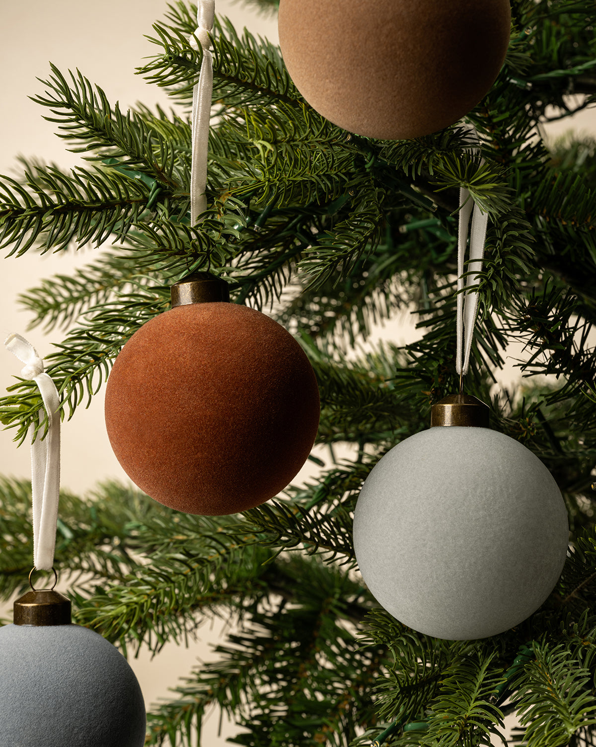 Close-up of a Christmas tree featuring the 3" Assorted Flocked Baubles (Set of 4) in brown, rust, and light gray, each ornament hanging from white ribbons among green pine needles, by Vendixs