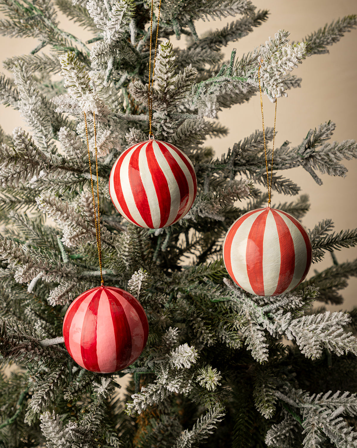 A set of three 3" hand-painted paper mache baubles with tassels hangs on a snow-frosted Christmas tree, adding a festive holiday touch, by Vendixs