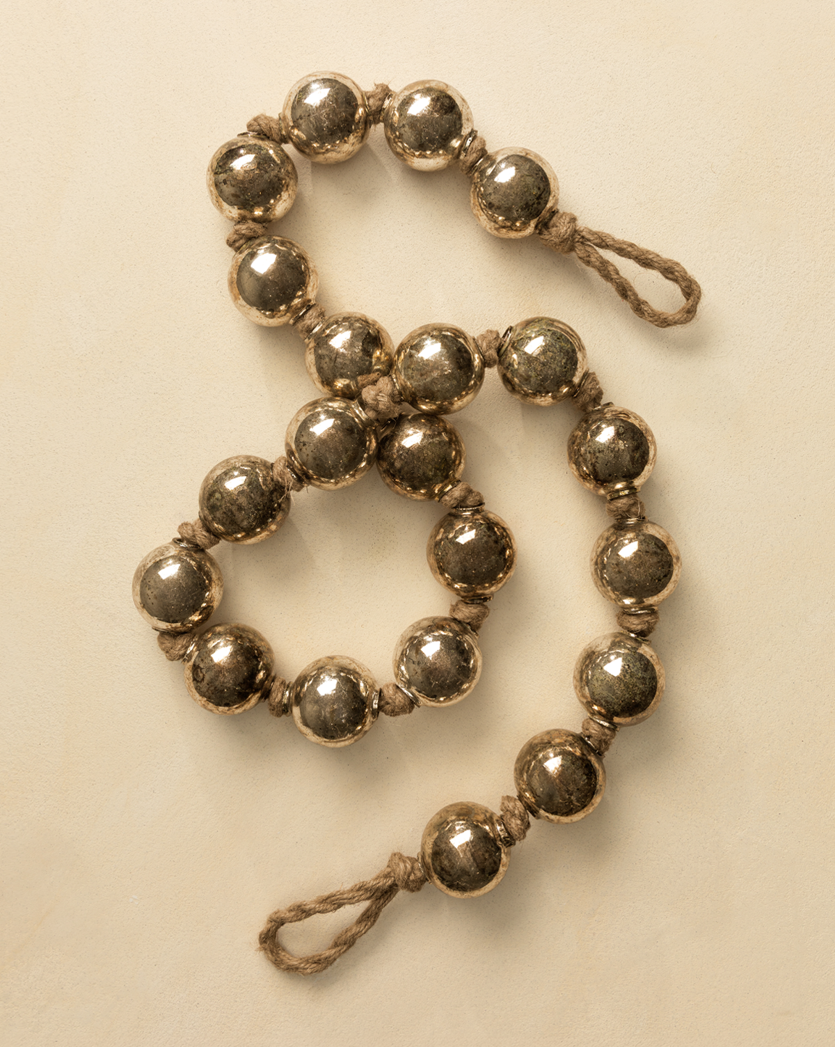 The 4.4' Aged Silver Glass Garland features large silver beads on thick jute twine with loops at each end, evoking vintage holiday decor and shown on a light beige surface, by Vendixs