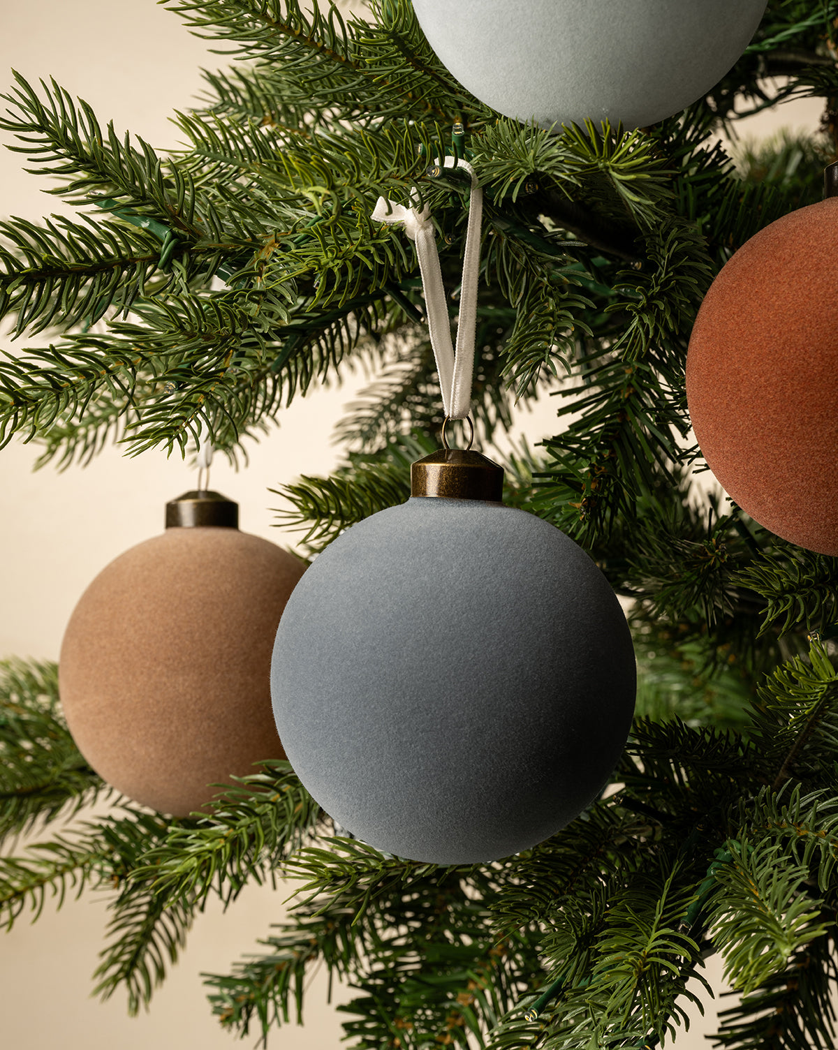 A close-up of a Christmas tree adorned with 4" Assorted Flocked Baubles (Set of 4) in matte blue, brown, and rust, adding festive flair to its green branches, by Vendixs