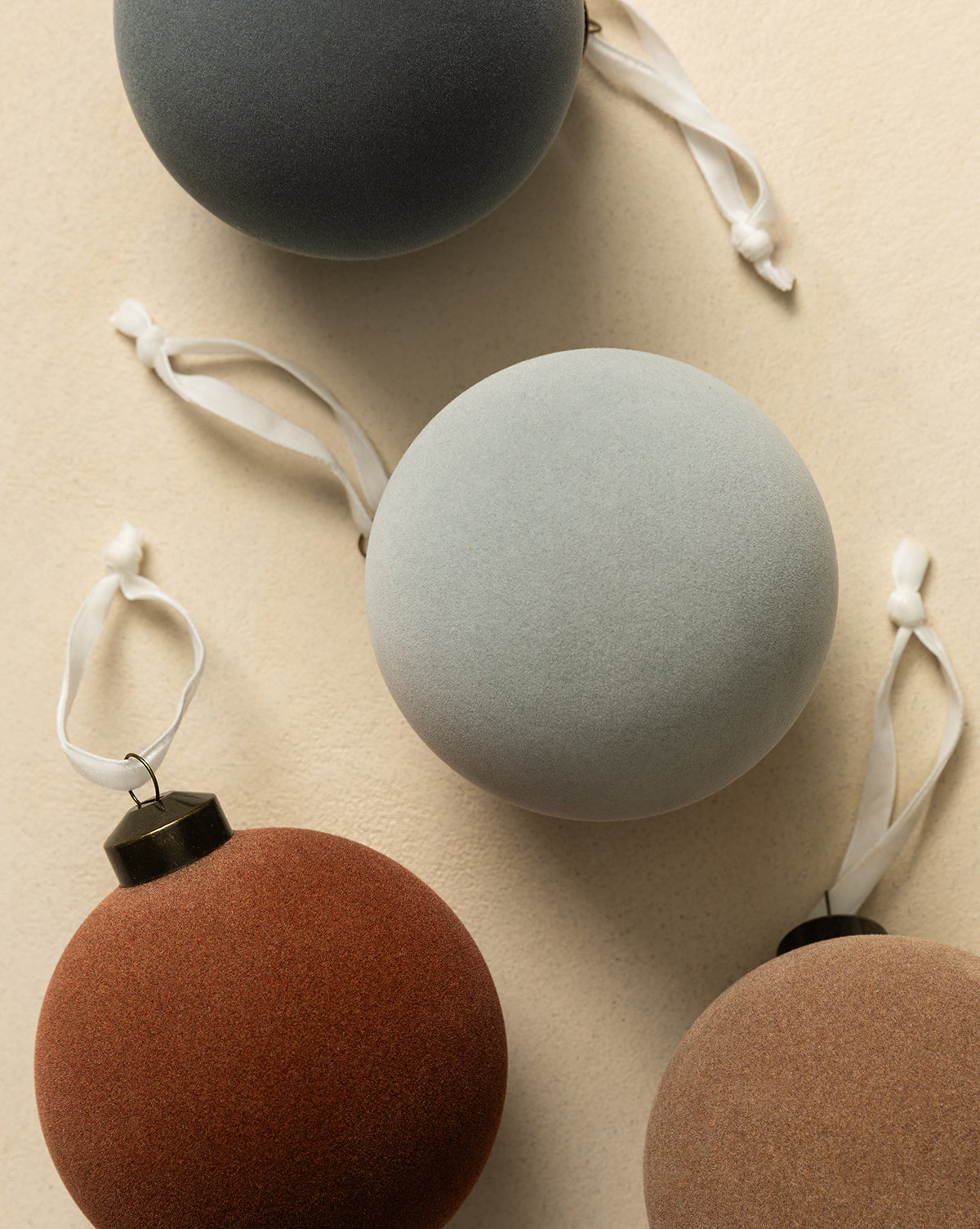 The 4" Assorted Flocked Baubles (Set of 4) features velvet-textured ornaments in gray, light blue, rust, and taupe with white hanging ribbons, perfect for adding an elegant touch to your holiday décor, by Vendixs