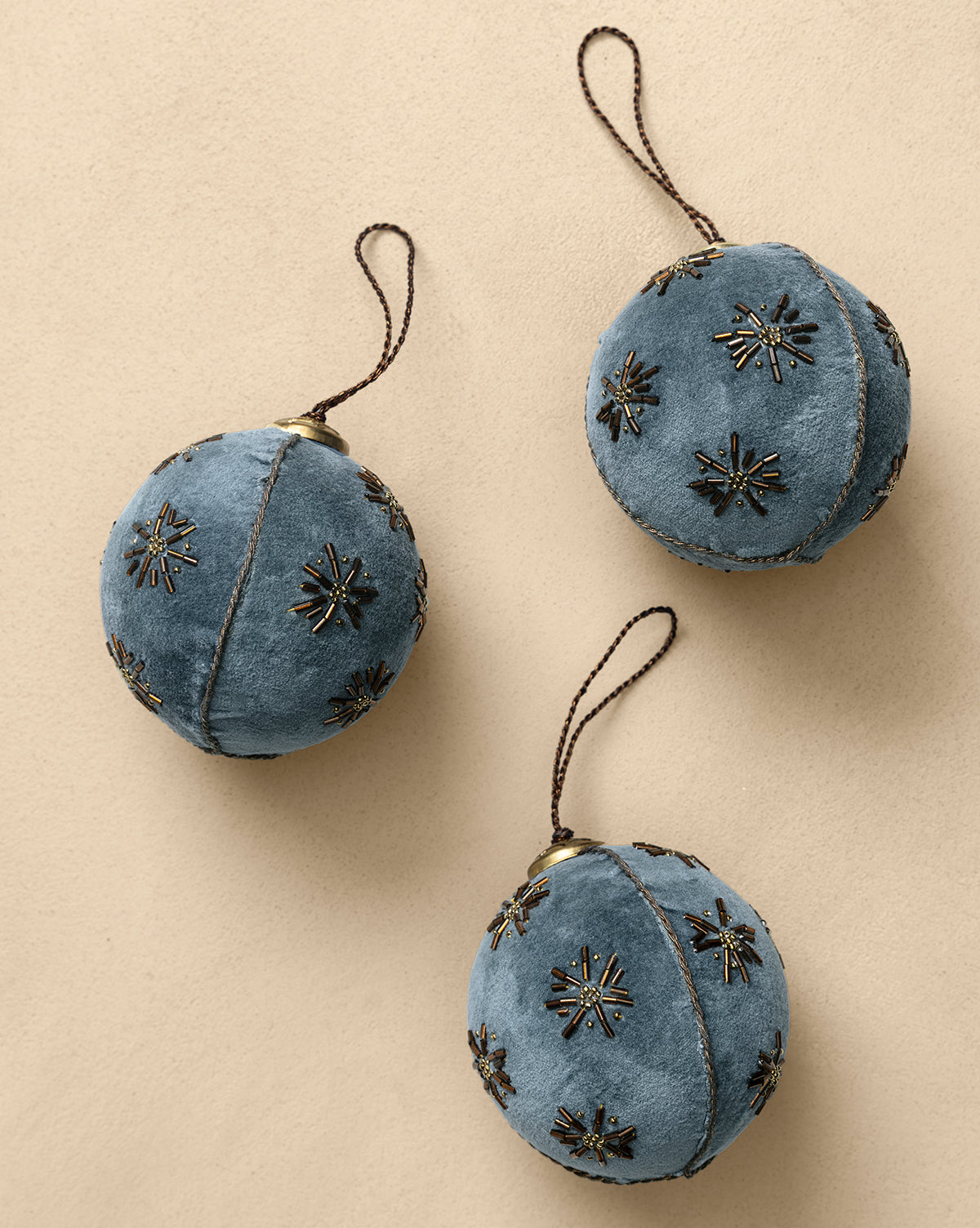 The 4" Embroidered Velvet Baubles (Set of 3) feature blue velvet with black and gold beaded starburst patterns and brown loops, bringing vintage-inspired elegance to any display on a beige background, by Vendixs
