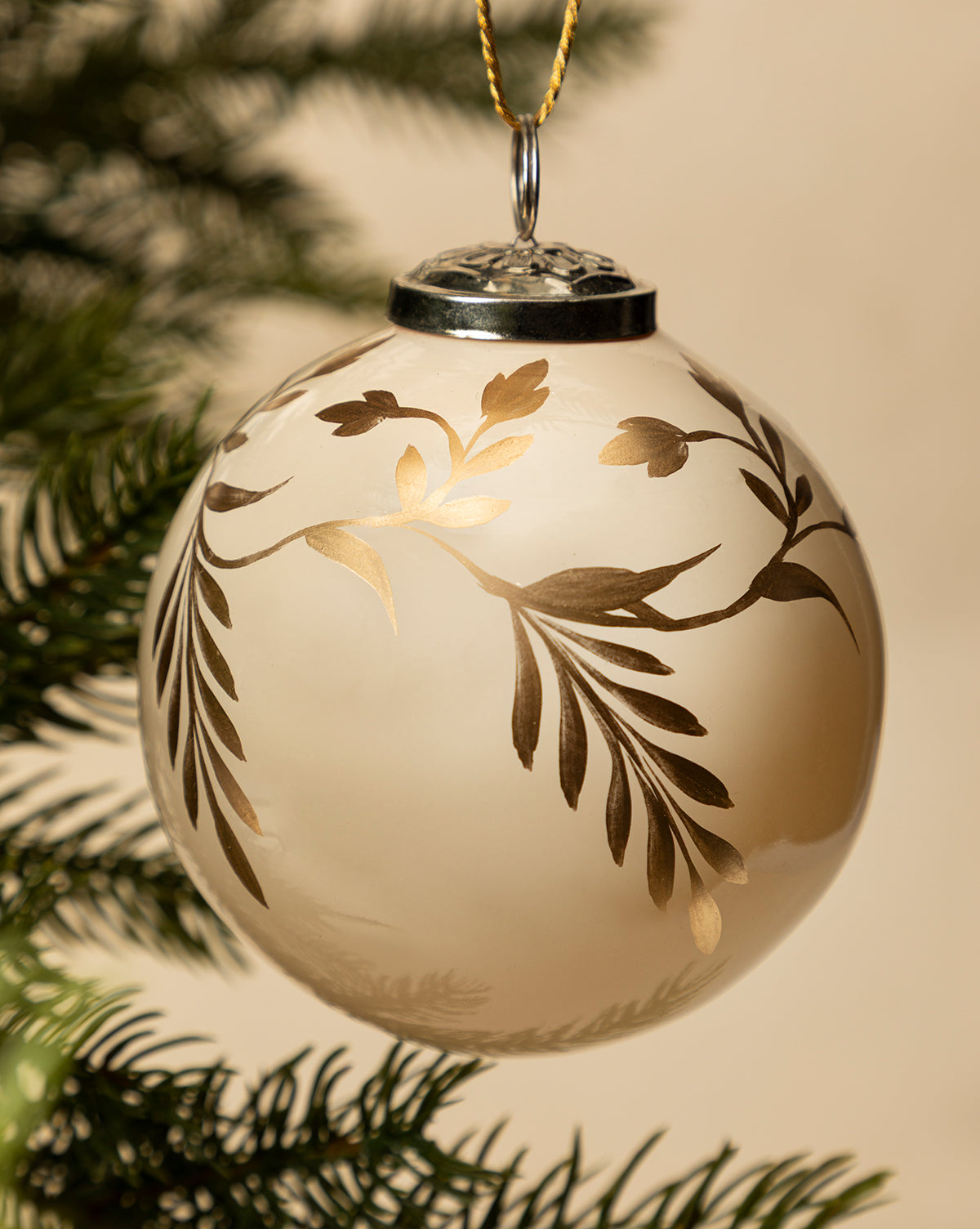 A close-up of the 4" Hand-Painted Vine Glass Bauble, featuring elegant gold leaf patterns, hangs from a pine branch—offering festive inspiration for holiday decorating with unique beaded ornaments, by Vendixs