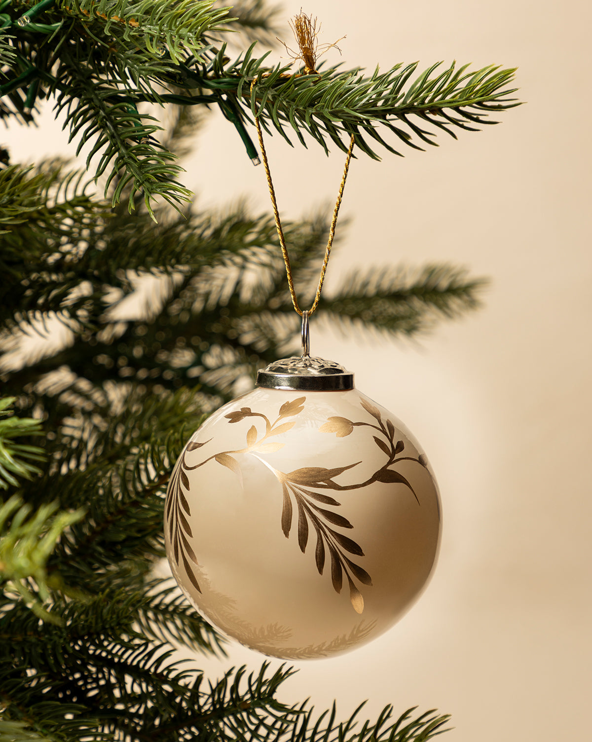The 4" Hand-Painted Vine Glass Bauble, featuring gold and white beads in a leafy design, hangs from a green pine branch and brings elegant style to your holiday decor against a neutral backdrop, by Vendixs