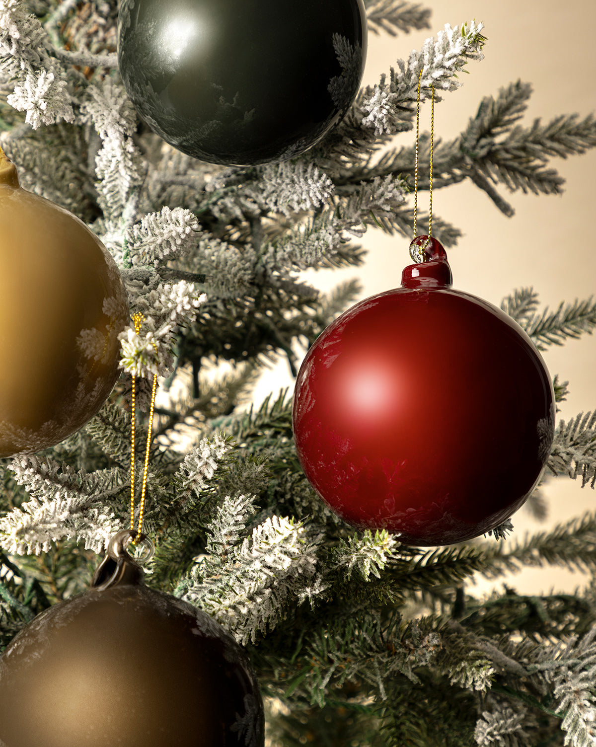Close-up of a Christmas tree with snow-dusted branches, decorated for the holidays with 4" High Gloss Glass Baubles (Set of 4) in red, green, and gold, hanging from shimmering gold strings, by Vendixs