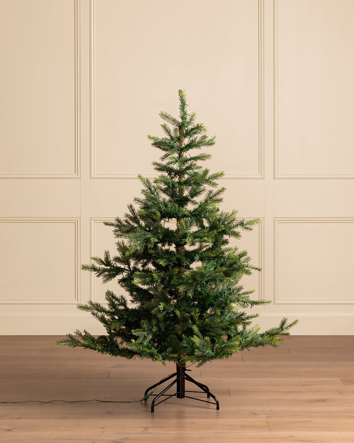 A 5' Grandis Fir Pre-Lit Faux Tree with sparse green branches stands on a metal base in a room with light wood floors and beige paneled walls, its cord indicating built-in lights for effortless holiday decorating, by Vendixs