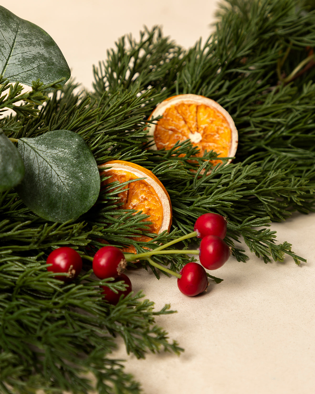 The 6' Orange & Eucalyptus Faux Garland, featuring green pine branches, red berries, large green leaves, and dried orange slices, adds eye-catching seasonal decor against a light beige background, by Vendixs