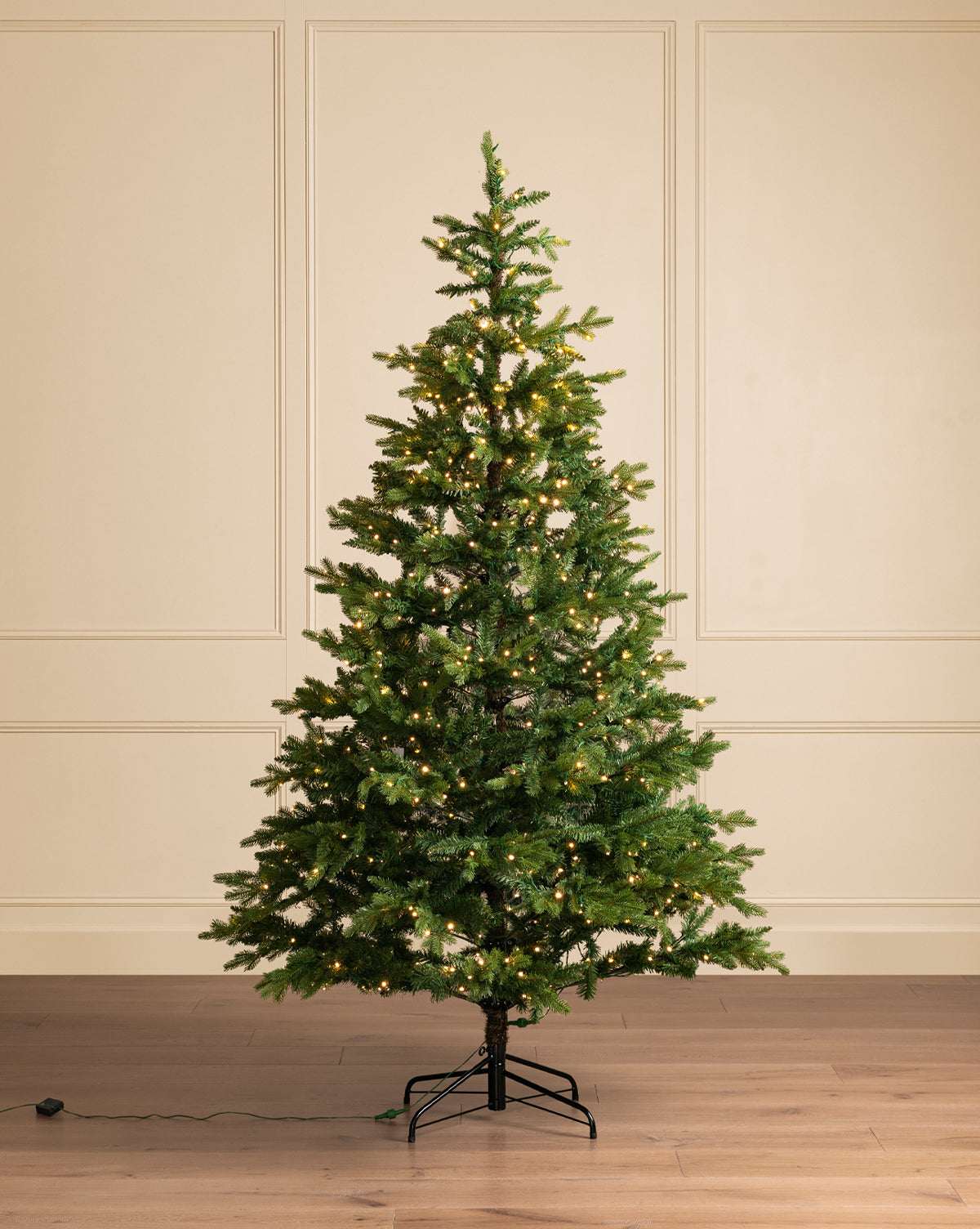 The 7' Allison Pine Pre-Lit Faux Tree stands on a metal base atop a wooden floor before a beige paneled wall, its warm white lights adding holiday charm. An electrical cord lies nearby, by Vendixs