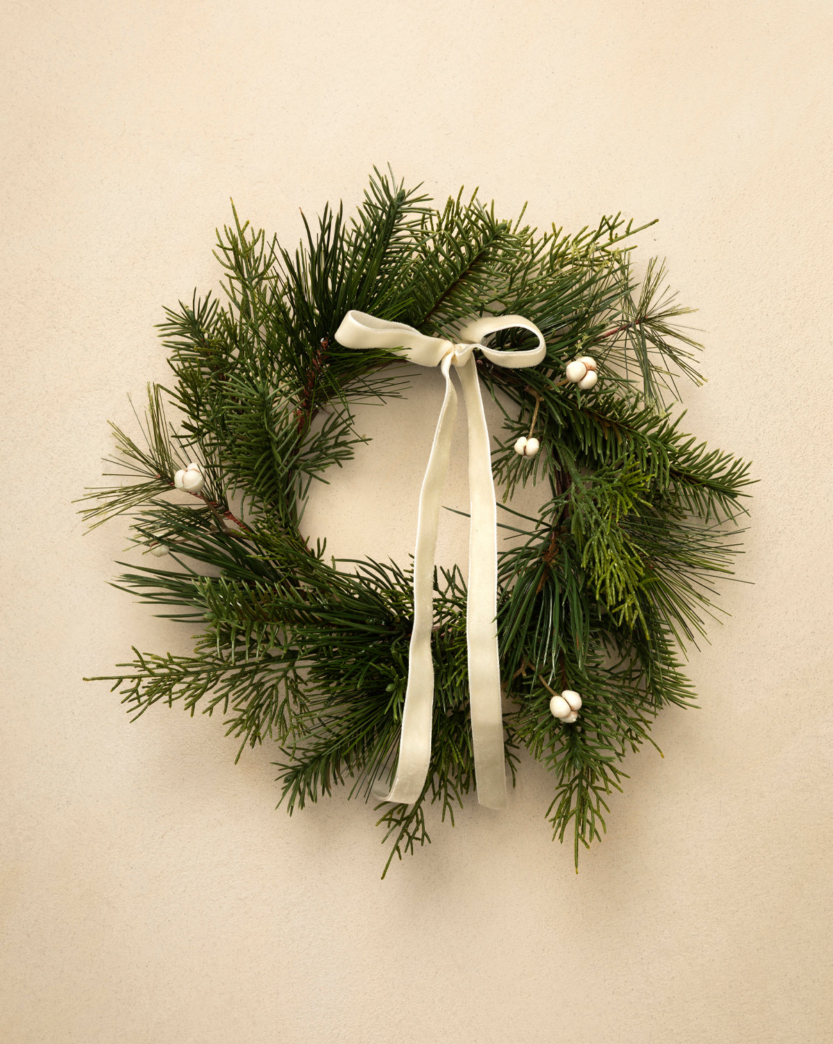 The 8" Pine Needle & Berry Faux Wreath, featuring small white berries and a white ribbon bow, adds a festive touch as it hangs on a light beige wall—ideal for holiday home decor, by Vendixs
