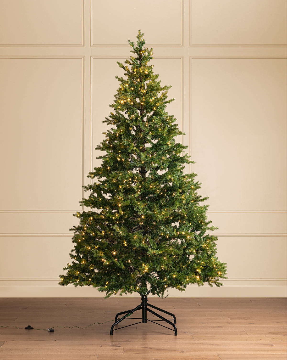 The 9' Allison Pine Pre-Lit Faux Tree, featuring realistic branches and warm white lights, is displayed on a metal base against a cream paneled wall and wood floor, by Vendixs