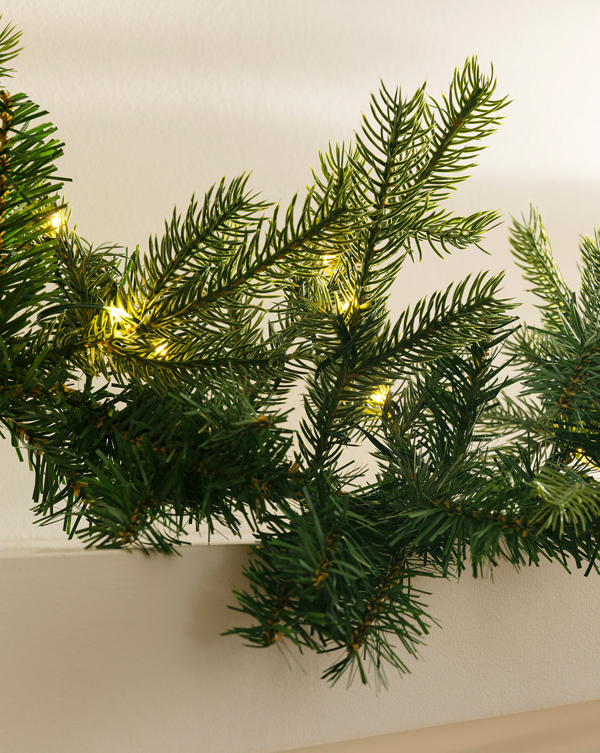 A close-up of the 9' Grandis Pre-Lit Garland showcases lifelike green pine branches with warm white lights, draped over a white surface—ideal for enhancing any indoor or outdoor holiday decor, by Vendixs