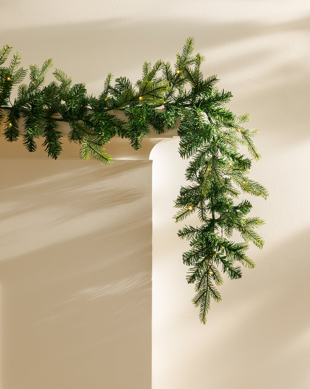 A 9' Grandis Pre-Lit Garland drapes over a white mantel, casting soft shadows in warm light—ideal for festive indoor or outdoor holiday décor, by Vendixs