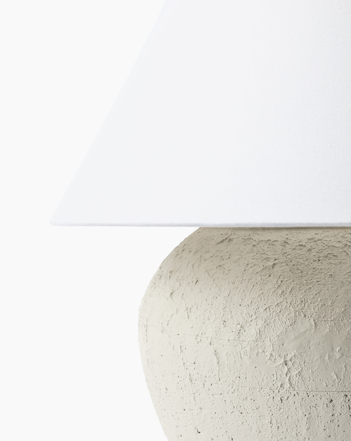 Close-up of the Abbott Ceramic Table Lamp, showcasing its textured, round ceramic base and the lower edge of a white linen shade against a plain white background, by Vendixs